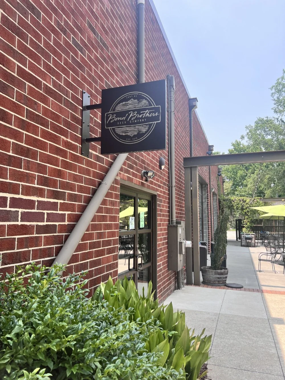 Exterior view of Bond Brothers Beer Company with a black sign on a red brick wall, outdoor seating area with tables and umbrellas, plants, and trees in the background.