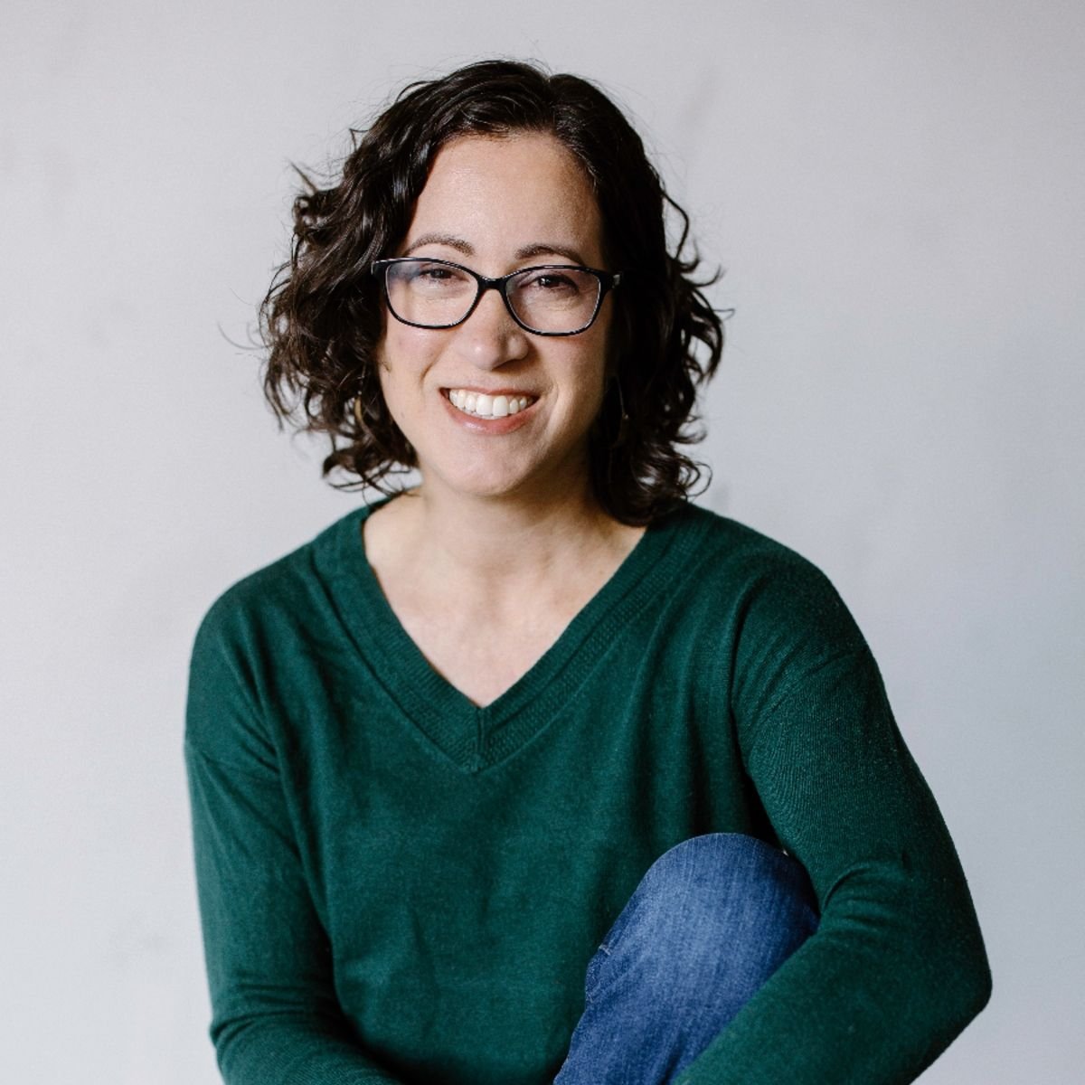 A woman with curly dark hair, glasses, and a green sweater smiling at the camera against a plain light background.
