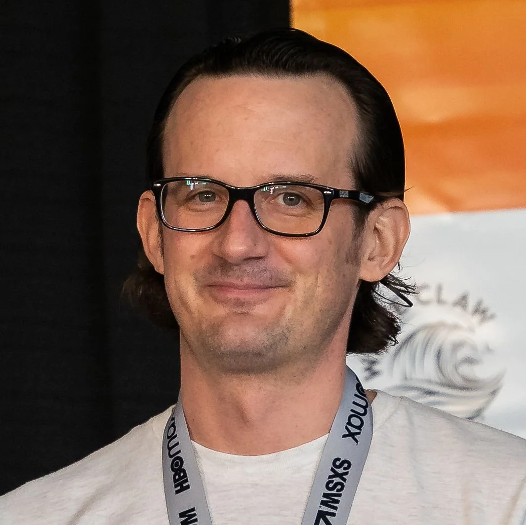 A man with dark hair, glasses, and a light beard, smiling in a casual setting.