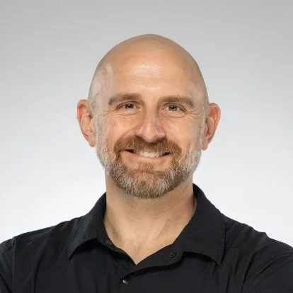Headshot of a man with a beard, smiling, wearing a black collared shirt, against a light gray background.