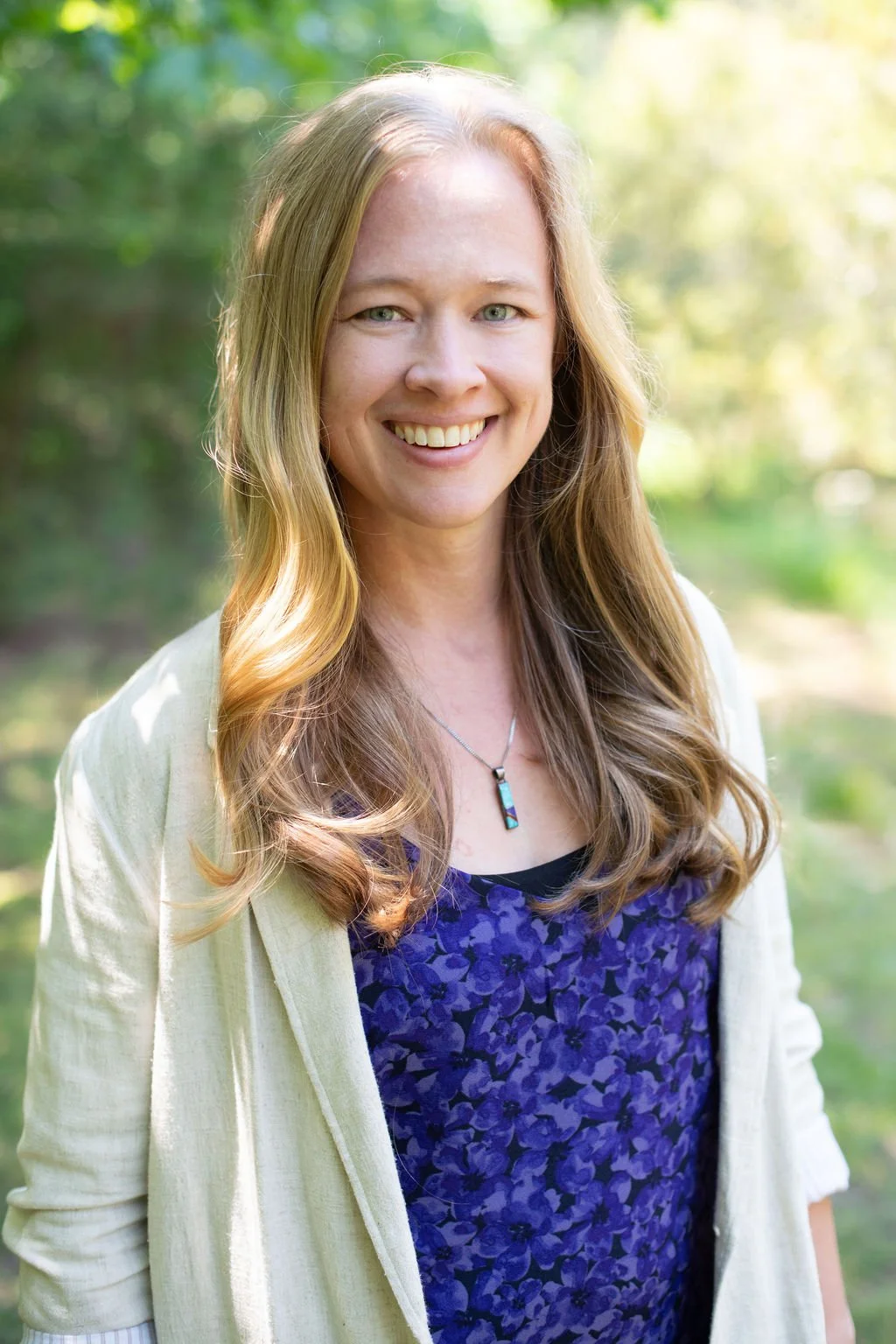 A woman with long, wavy blonde hair, smiling outdoors with a green, leafy background.