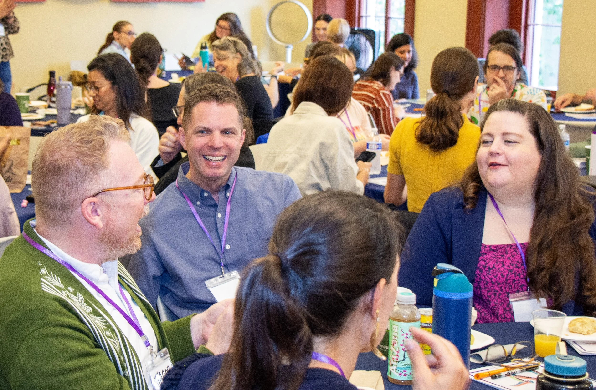 People sitting at tables, engaged in conversations, with food and drinks, in a bright, colorful room during a social or professional event.