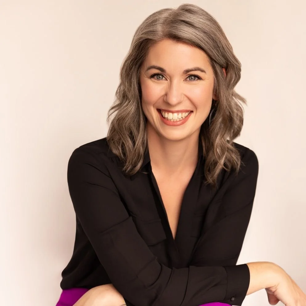 A woman with gray wavy hair smiling at the camera, wearing a black top and purple pants, sitting against a plain light-colored background.