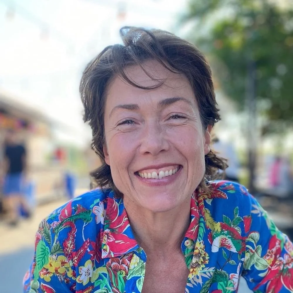 A woman smiling outdoors, wearing a colorful floral shirt, with trees and blurred people in the background.