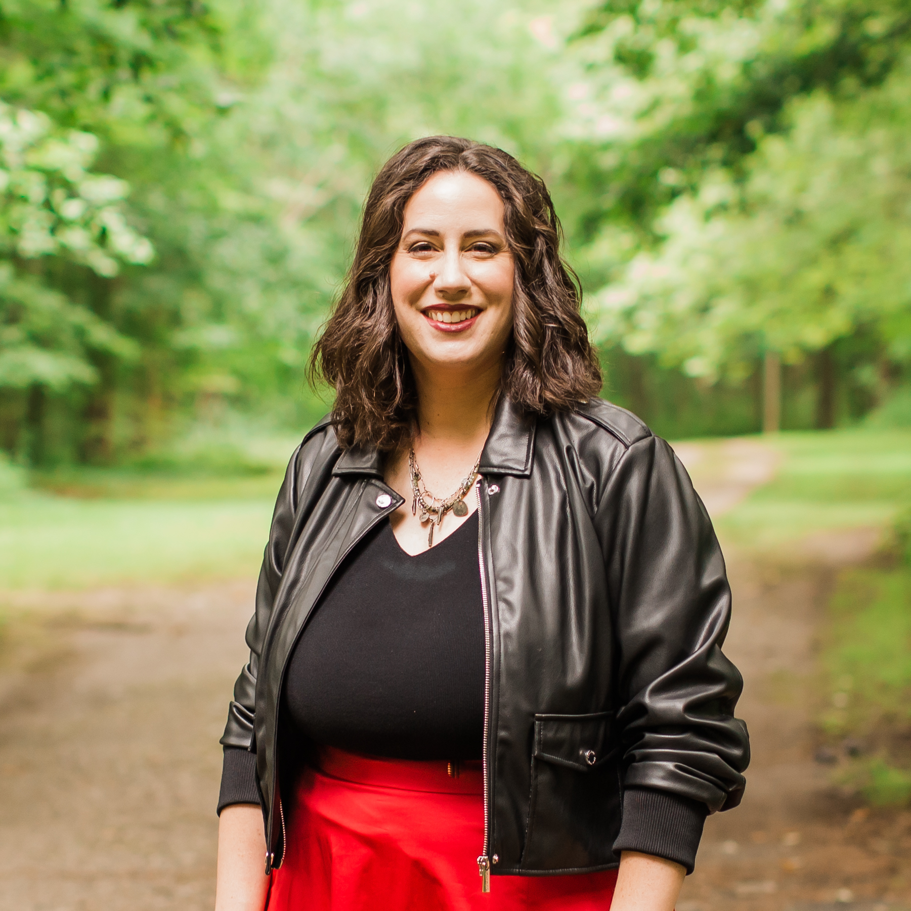 A woman with brown curly hair smiling outdoors in a park, wearing a black leather jacket over a black top and red skirt.