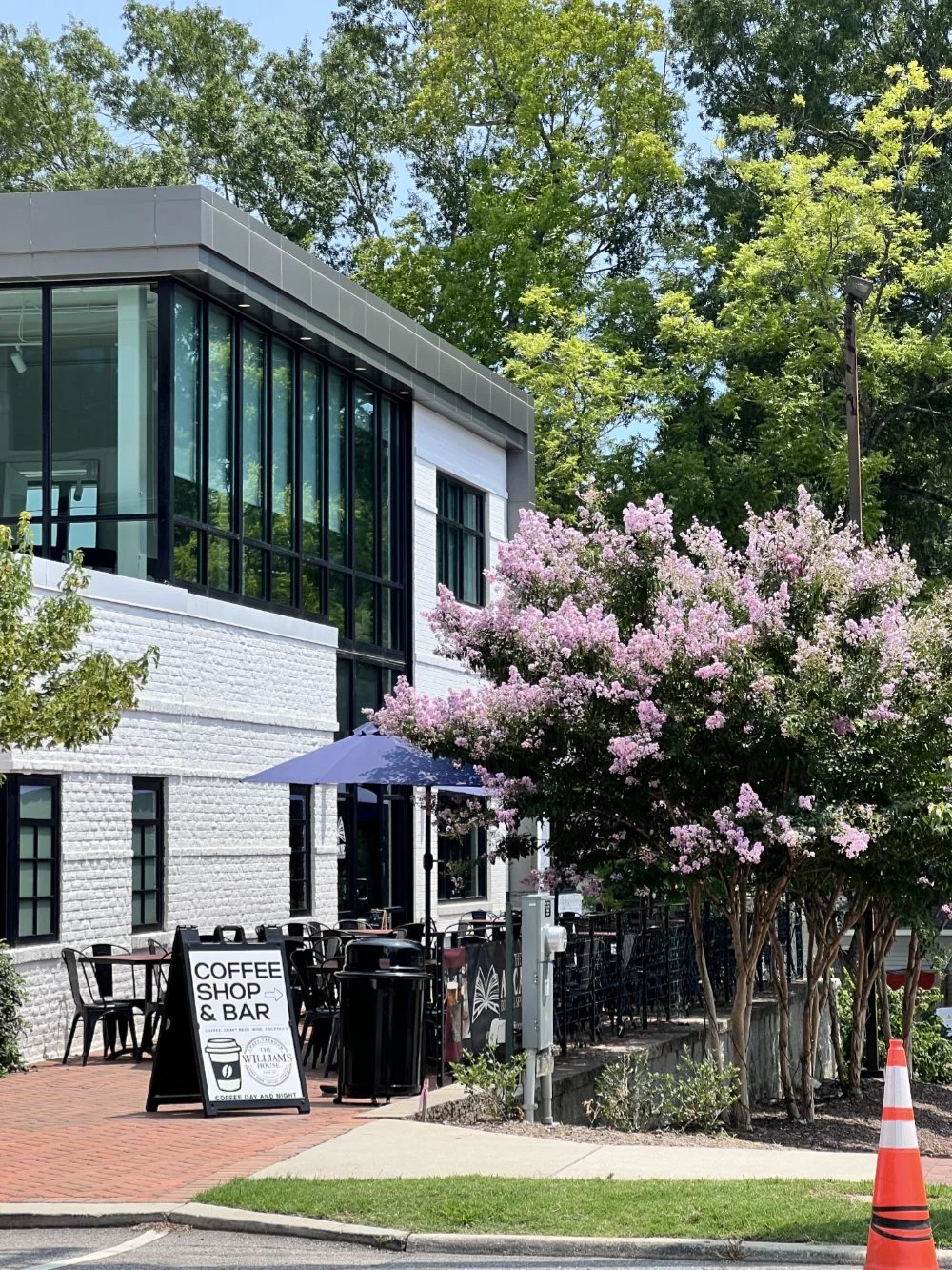 Exterior of a modern building with large glass windows, outdoor seating with chairs and a purple umbrella, a blooming pink flowering tree, and a sign for a coffee shop and bar.