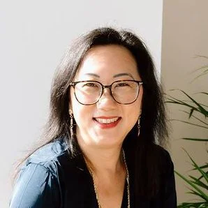 A woman with black hair, wearing glasses and earrings, smiling in a well-lit room with plants in the background.