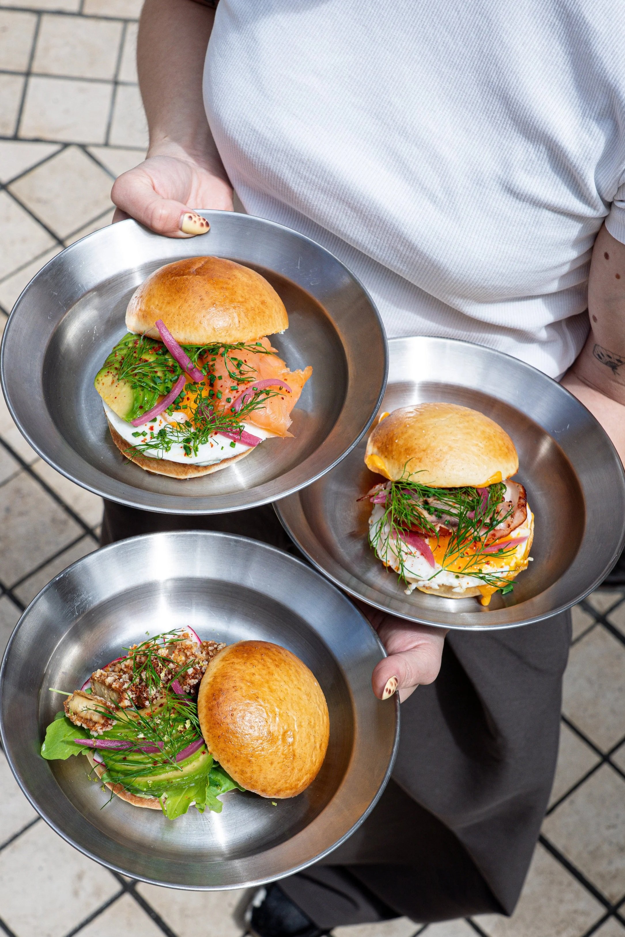 Deux personnes tenant des assiettes avec des sandwiches garnis de saumon fumé, de légumes, et d'herbes, sur une terrasse pavée.