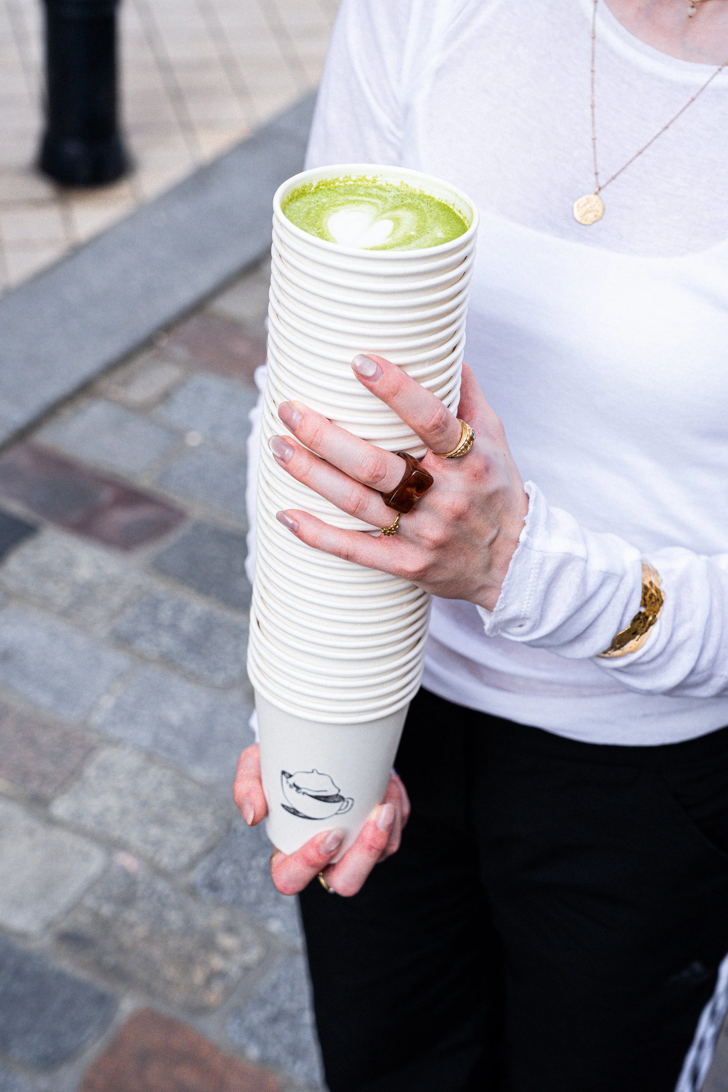 Une personne tient un grand gobelet blanc avec un matcha latte, en extérieur sur un pavement en pierre.