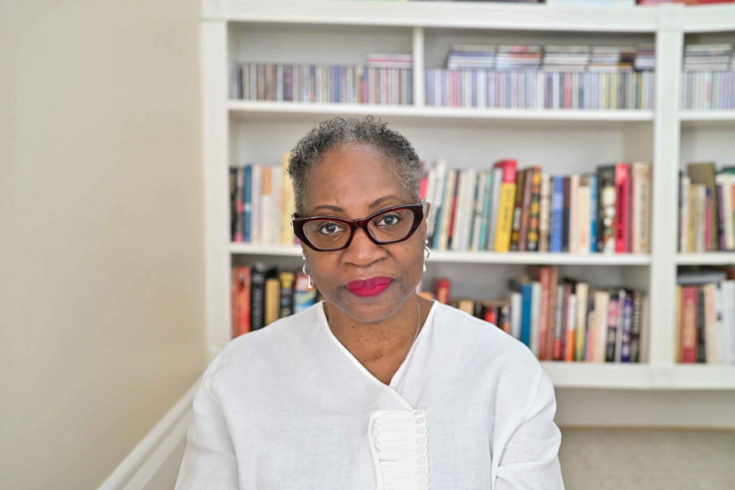 Dr. Claudia Mathis with short gray and black curly hair, wearing glasses and a white top, standing in front of a white bookshelf filled with colorful books.