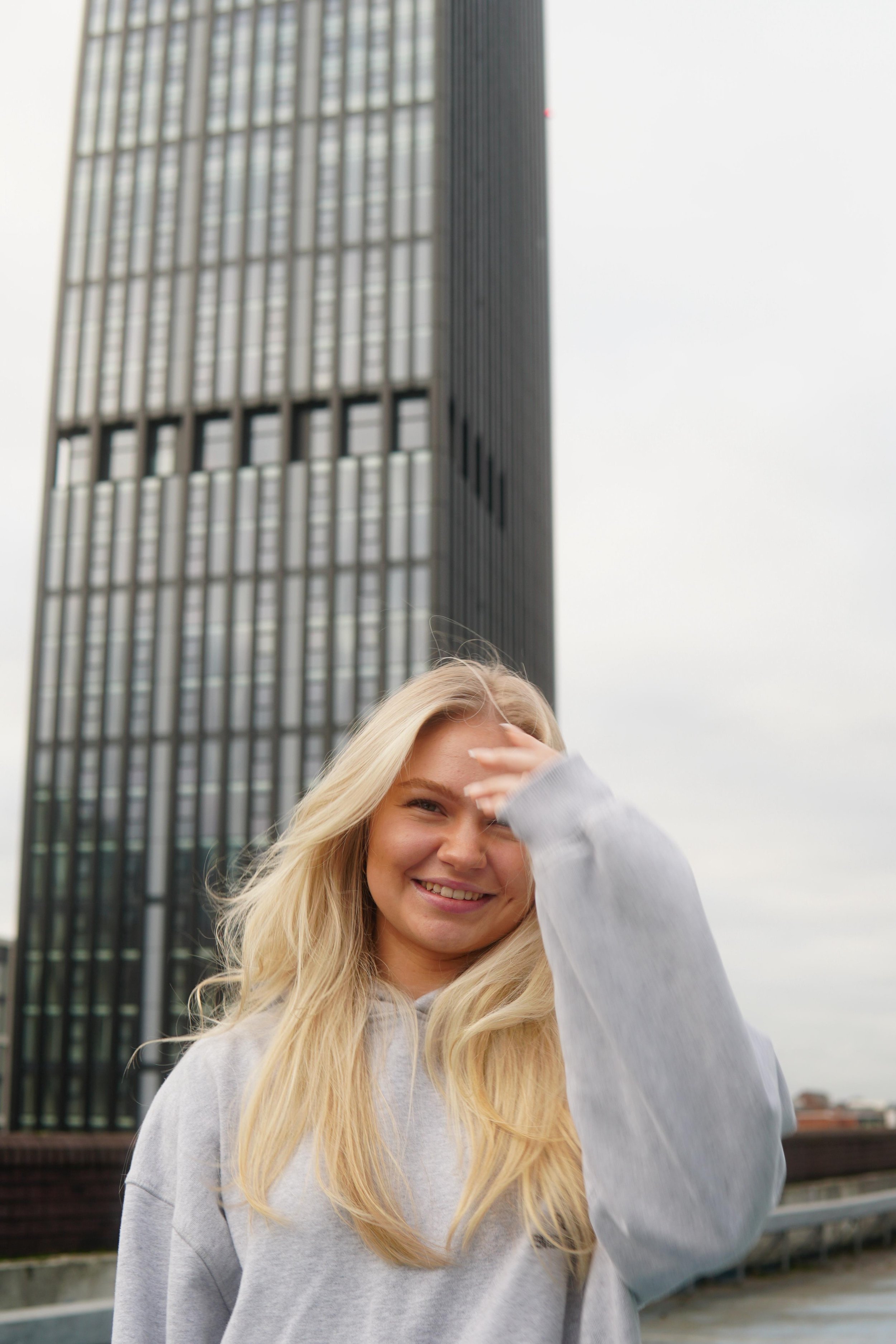 Young woman smiling and adjusting her hair outdoors with a modern skyscraper in the background.