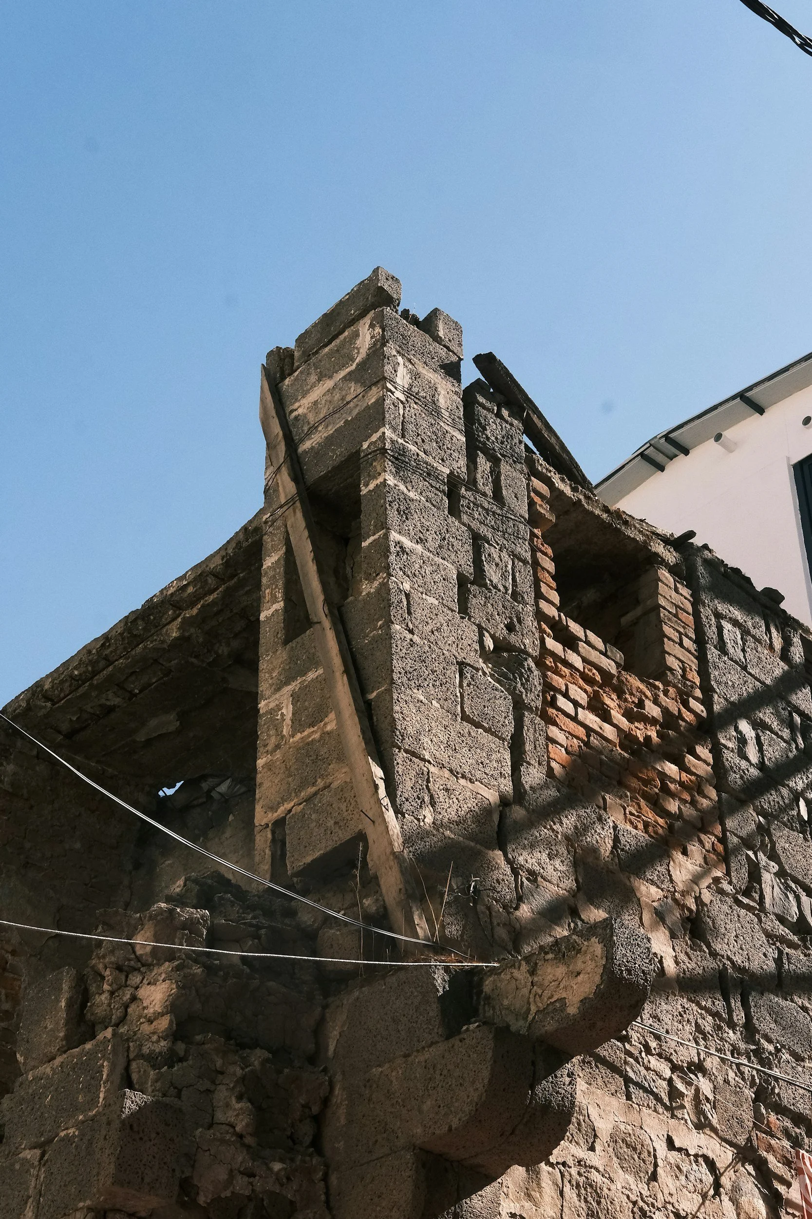 A damaged building with a partially collapsed brick wall and rubble at the base, under a clear blue sky.