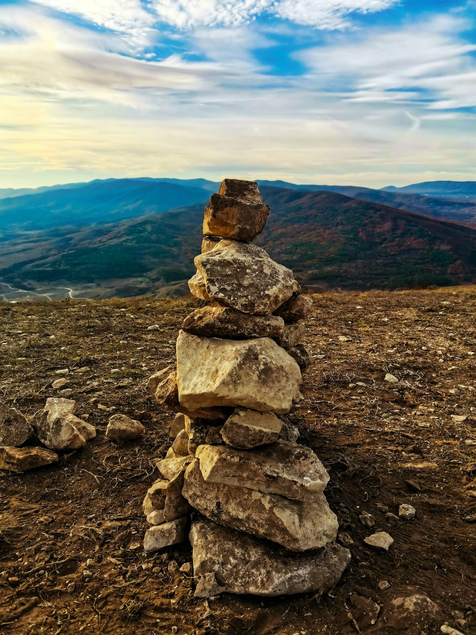 Stack of rocks arranged in a cairn on a dirt surface with mountain ranges and a cloudy sky in the background.