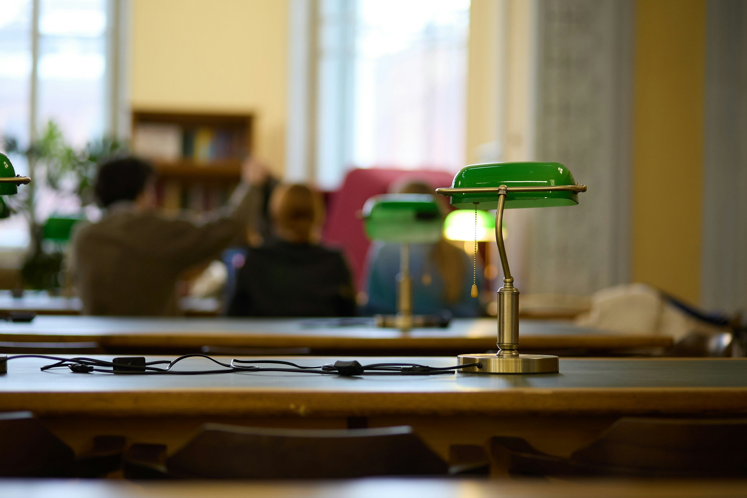 A green banker's desk lamp on a wooden table in a library or study room with blurred background of windows and a person reading.
