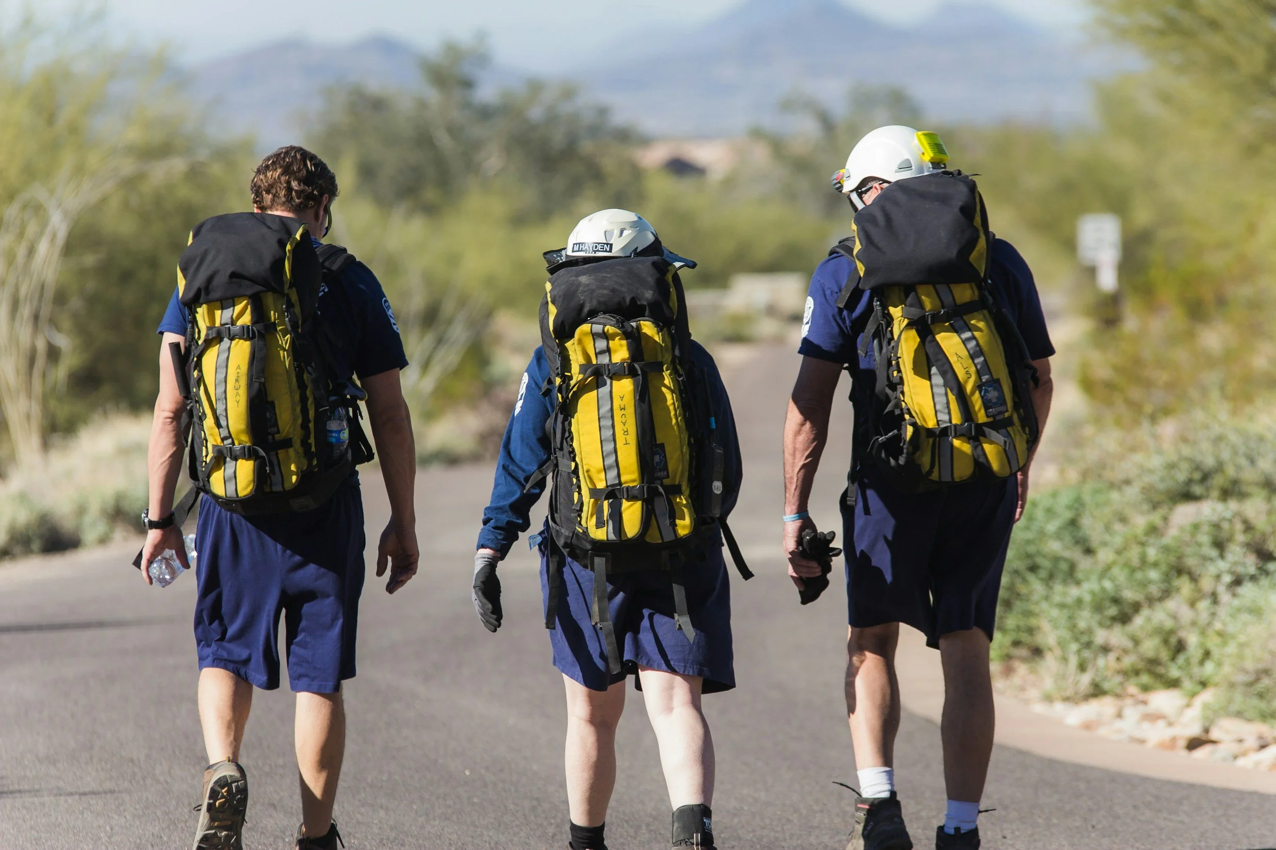 Four people in athletic clothing and helmets walking on a paved trail outdoors with greenery and mountains in the background.