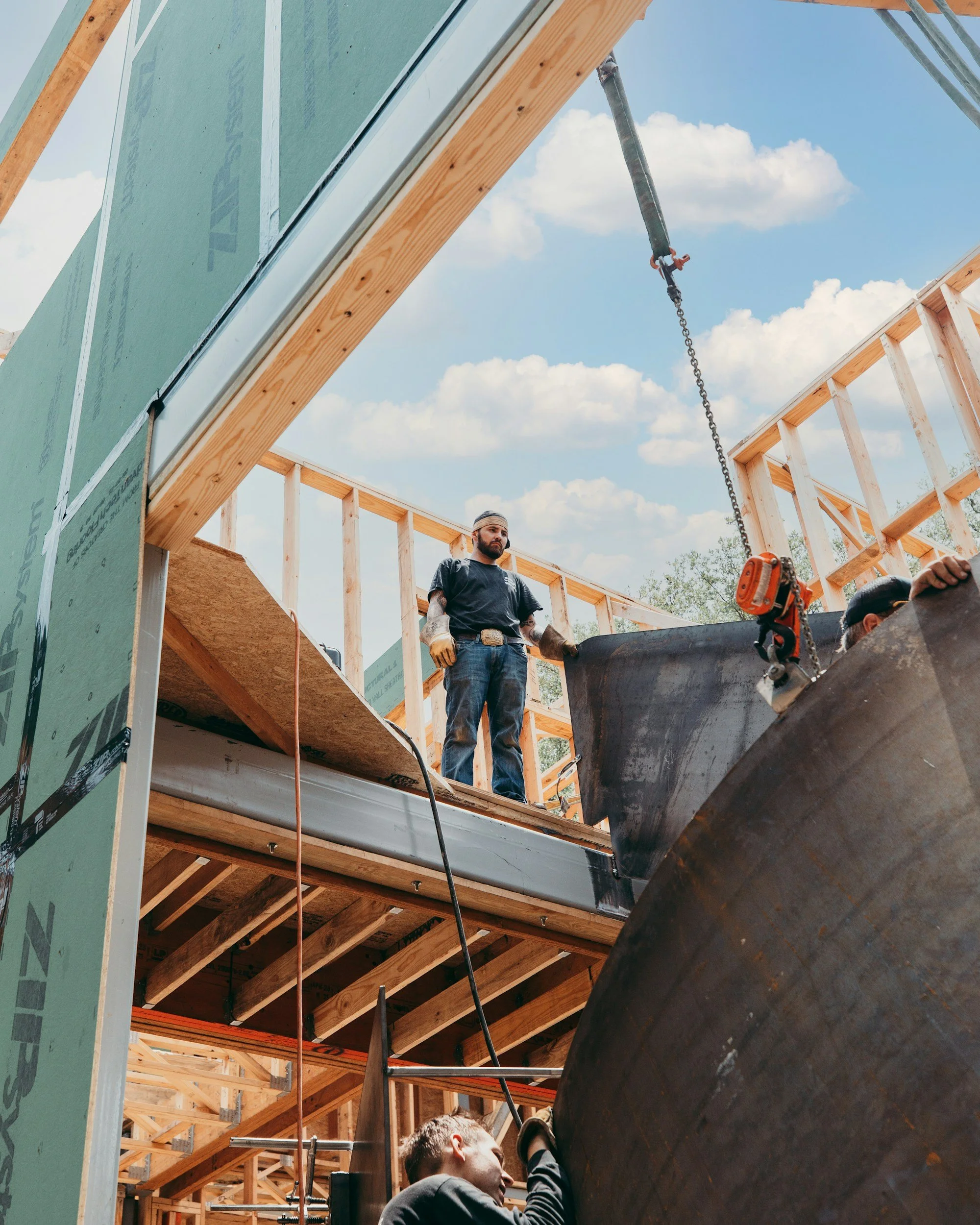 Construction workers installing a large metal pipe in a building under construction, with wooden framing and clear sky visible outside.