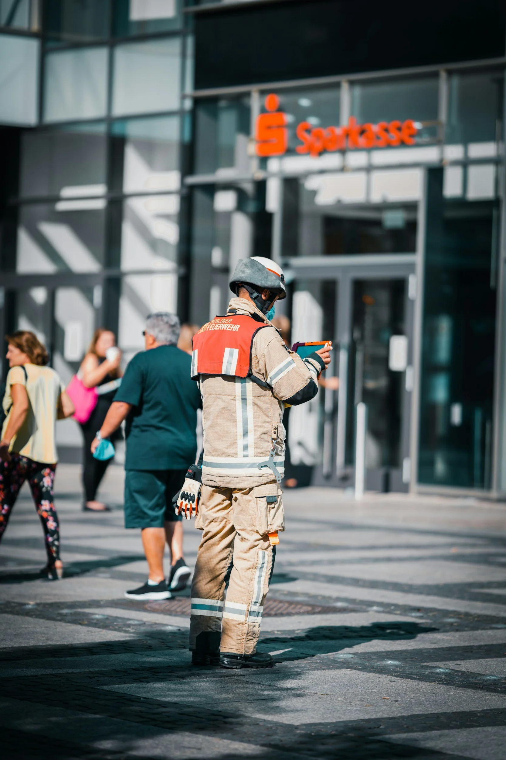 A firefighter in beige protective gear and helmet is standing in a crosswalk on a city street, looking at a device. People walk past behind her, and a modern glass building with an illuminated red and orange sign is in the background.