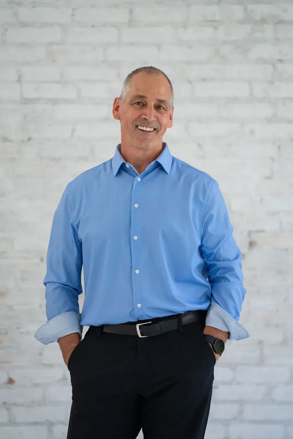Jim Brayshaw - A middle-aged man with short gray hair, wearing a light blue dress shirt and black pants, smiling and standing against a white brick wall.