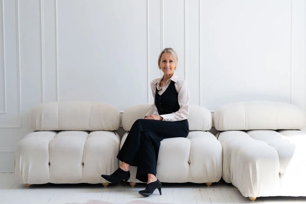 Angellah Brayshaw - A smiling woman sitting on a white sofa in a bright room.