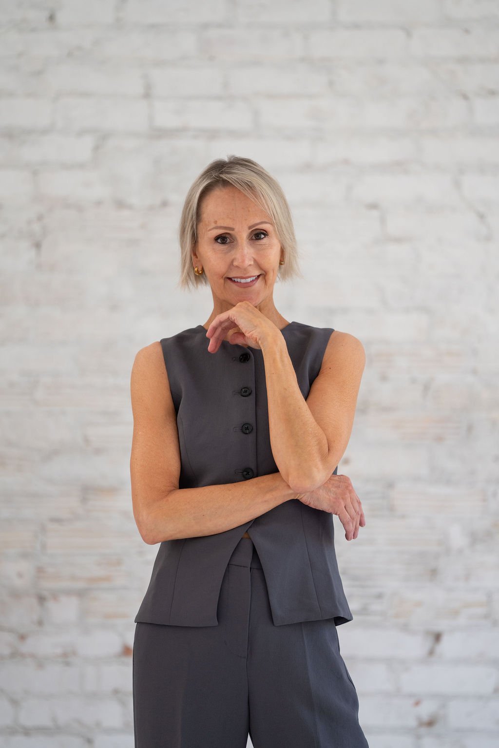 Angellah Brayshaw - A middle-aged woman with short blonde hair, wearing a sleeveless gray dress with black buttons, smiling and posing with her hand near her chin, against a white brick wall background.