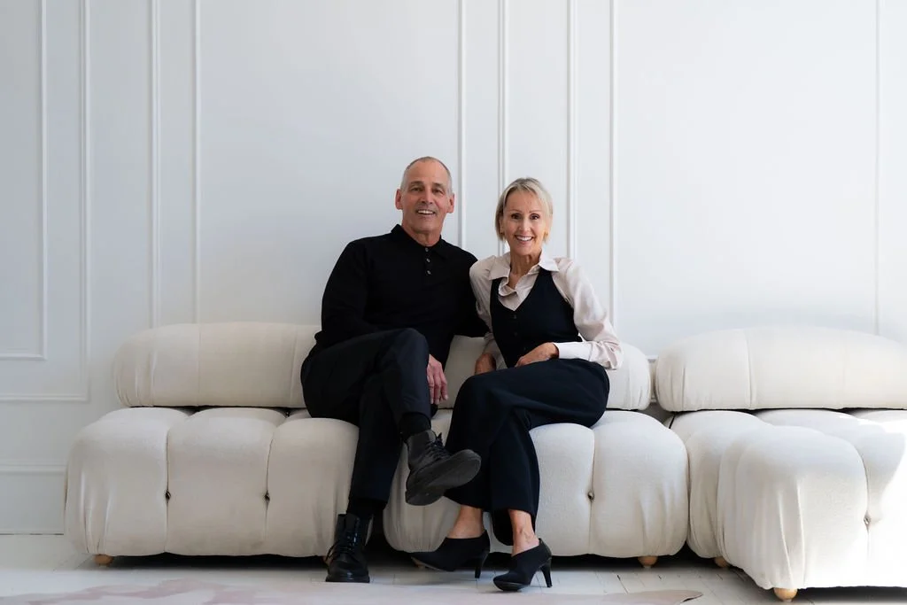 Jim & Angellah Brayshaw - A smiling older man and woman sitting close together on a white sofa in a bright, modern room with white walls.