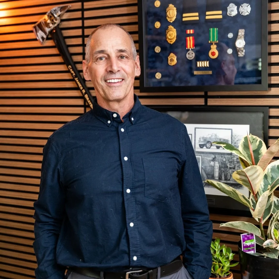 Jim Brayshaw - Man wearing a dark blue button-up shirt standing indoors in front of a wooden wall with military medals display and framed photographs, including a plant in the foreground.