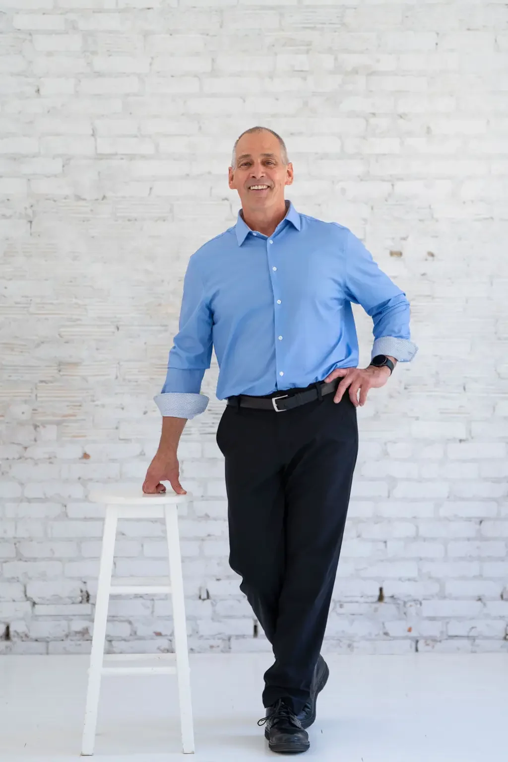 A man in a blue dress shirt and black pants standing next to a white stool against a white brick wall, smiling and posing with one hand on his hip.