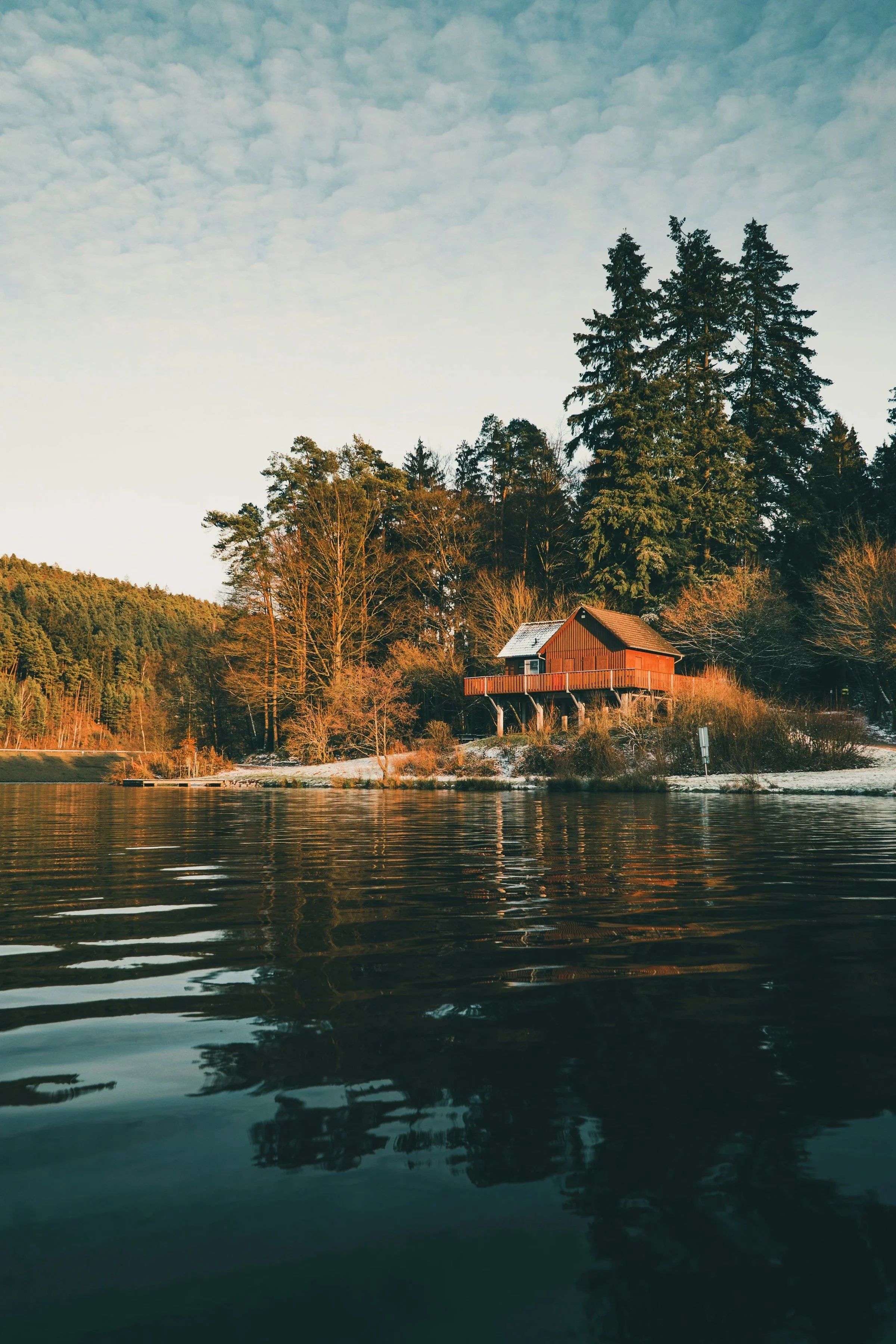 A house with a red exterior and a small porch is located on a hill beside a body of water. Tall trees and dense forest surround the house, with a partly cloudy sky overhead.