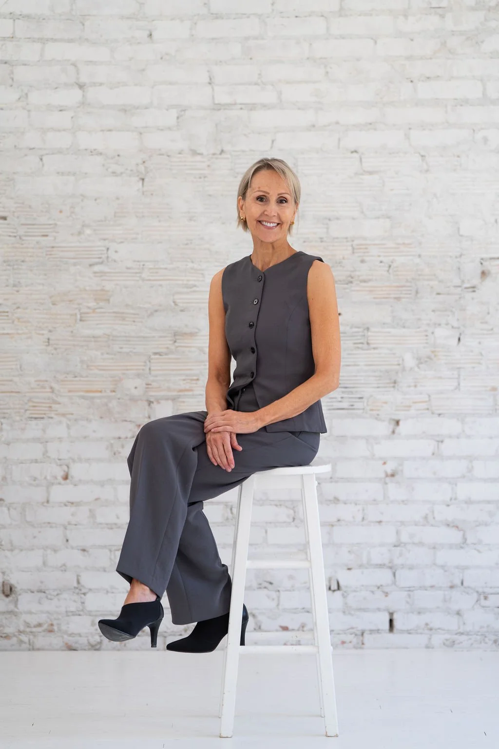 Smiling woman in gray sleeveless top and pants, sitting on a white stool against a white brick wall.