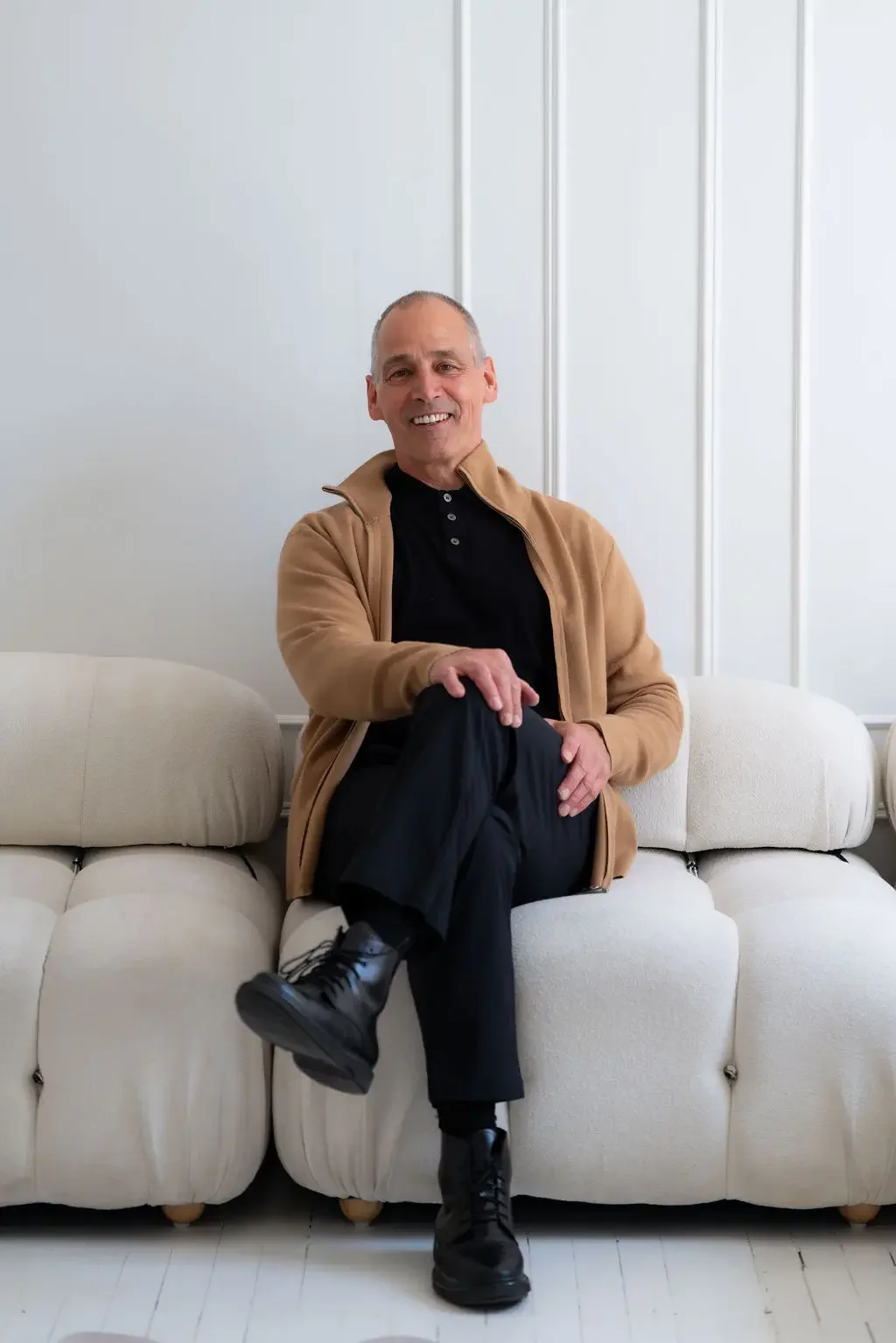 A smiling man with short gray hair sitting on a beige sofa in a white room, wearing a black shirt, a tan jacket, black pants, and black boots, with one leg crossed over the other.