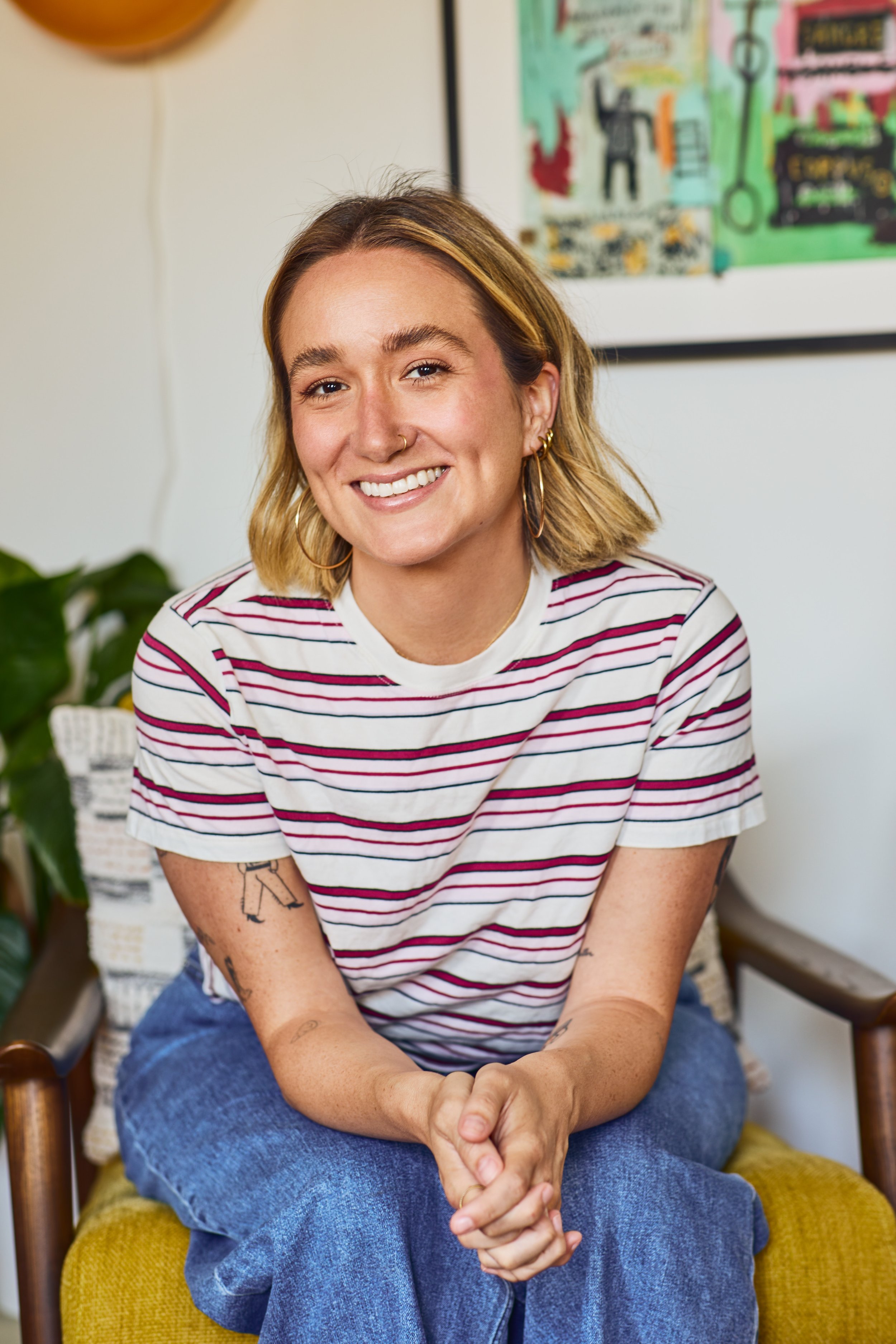 A smiling young woman with shoulder-length blonde hair, wearing a white and red striped t-shirt and blue jeans, sitting on a yellow chair in a cozy room with wall art and a potted plant in the background.