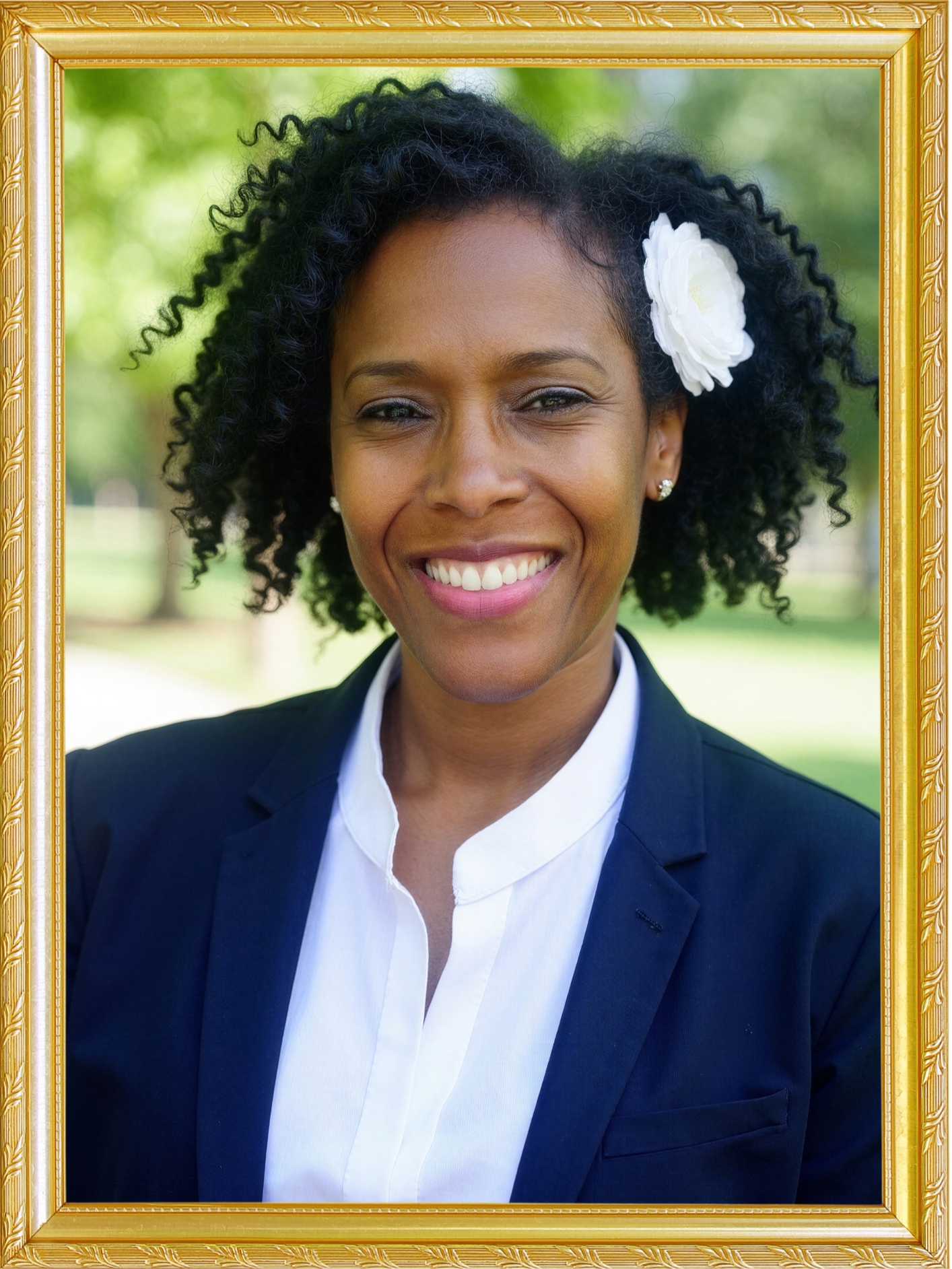 A smiling African American woman with curly black hair wearing a white flower hair accessory, a dark suit, and a white shirt, standing outdoors with a blurred green background, framed in a gold ornate picture frame.