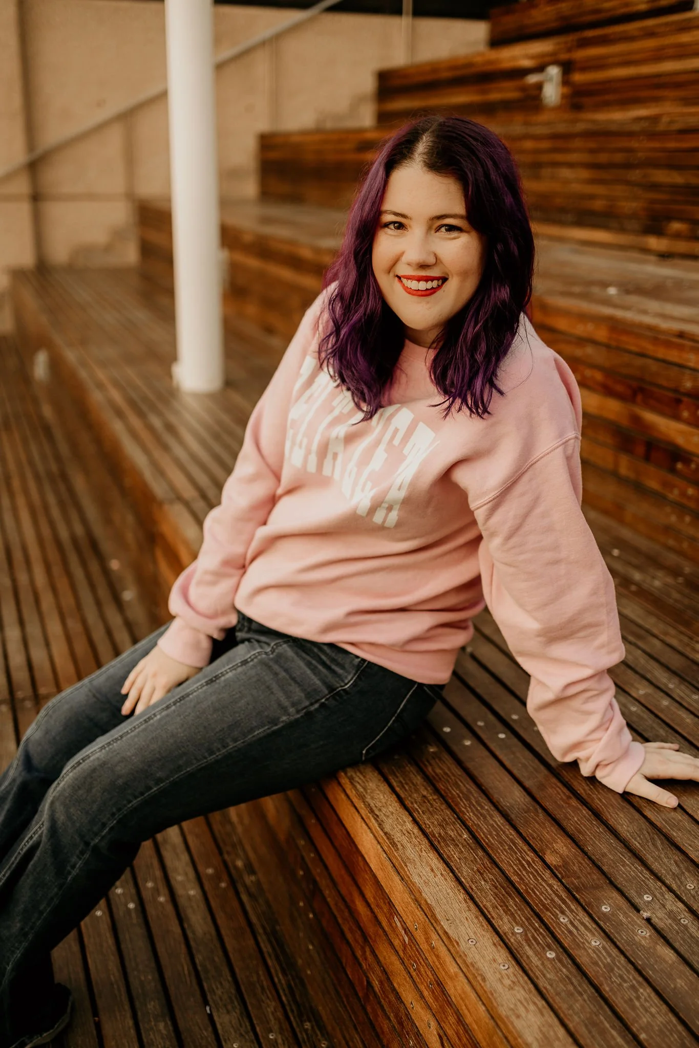 A young woman with purple hair, wearing a pink sweatshirt and dark jeans, sitting on a wooden bench, smiling at the camera.