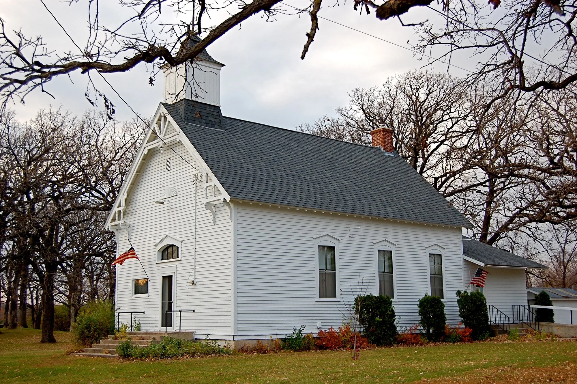White church with a black shingled roof, brick chimney, and steeple surrounded by leafless trees under a cloudy sky.