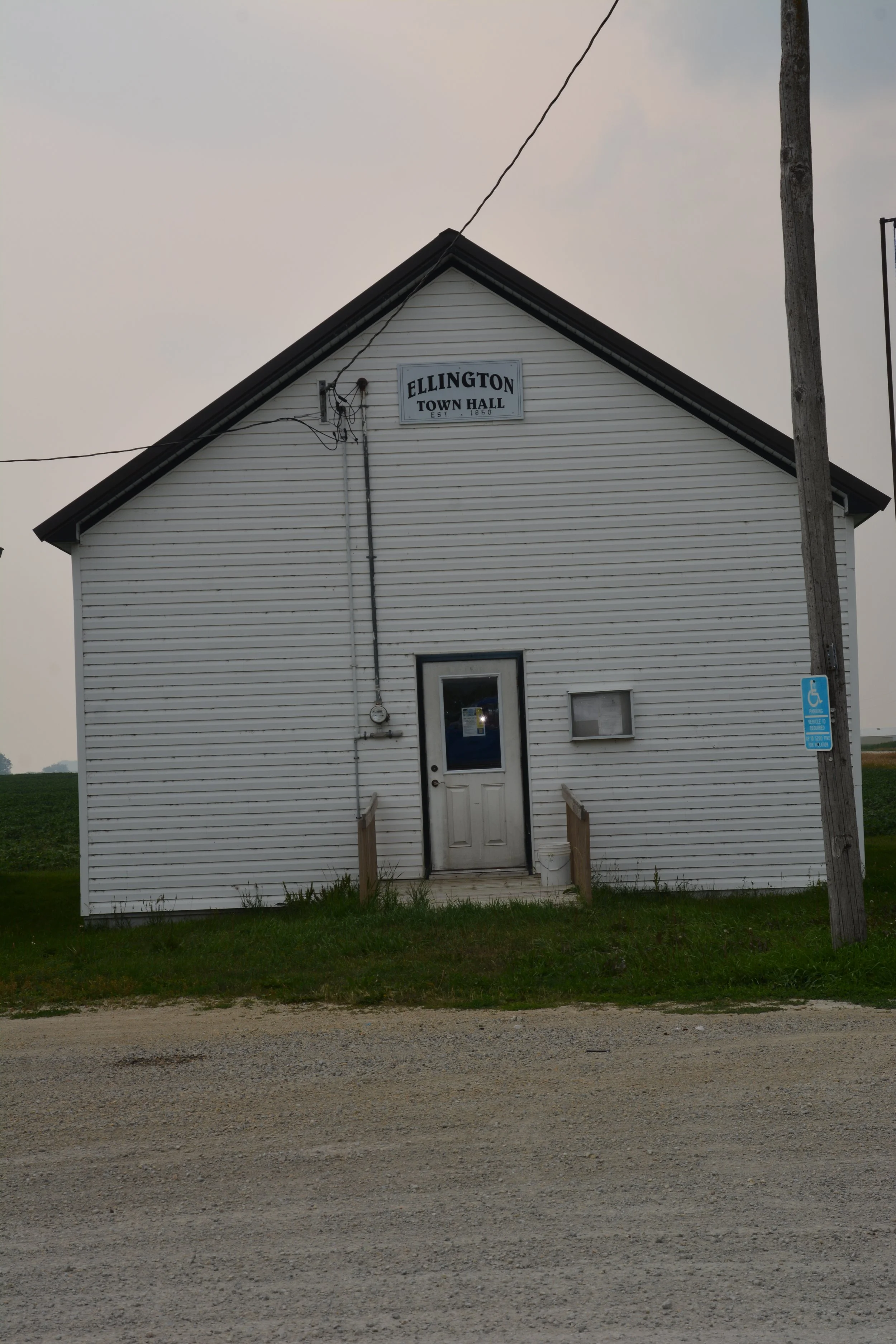 White building with a sign that reads 'Ellington Town Hall Established 1890', with two small steps leading to the door, a handicap parking sign on a pole to the right, and a utility pole with wires in front.
