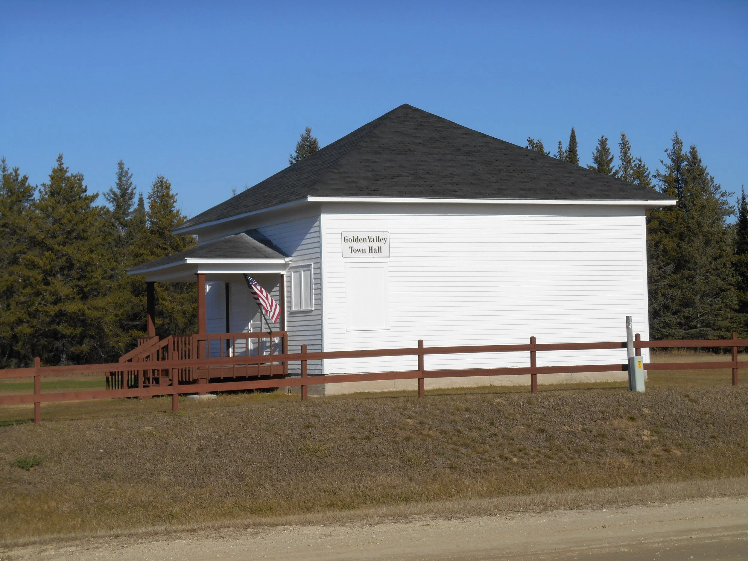 White town hall building with a dark gray roof and a porch, surrounded by a brown wooden fence, with trees in the background and a clear blue sky.