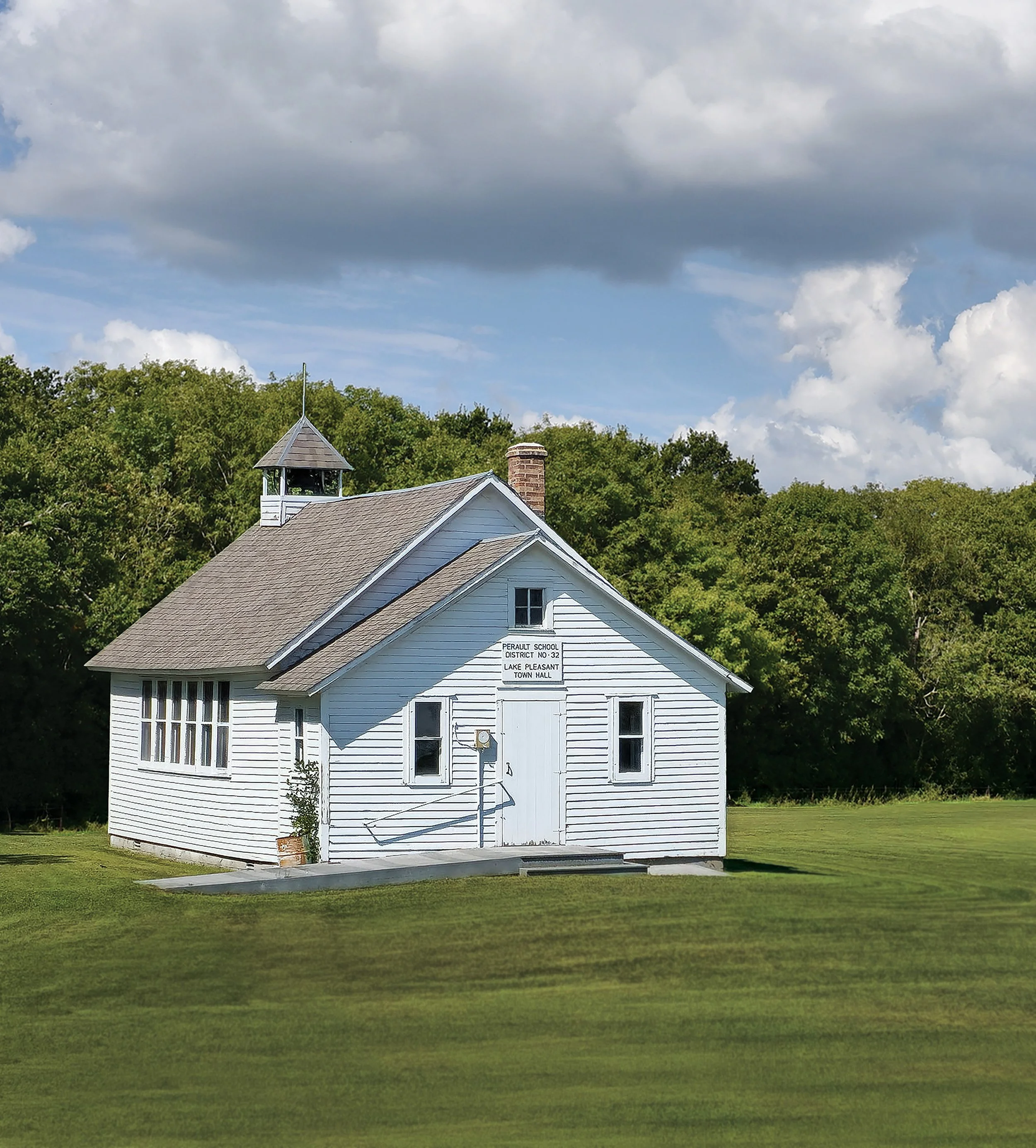 White wooden building with a pointed roof and a small bell tower on top, surrounded by green trees and grass, under a cloudy sky, with a sign that reads 'PERALUIT SCHOOL DISTRICT NO. 32 LAKE PLEASANT TOWN HALL'.