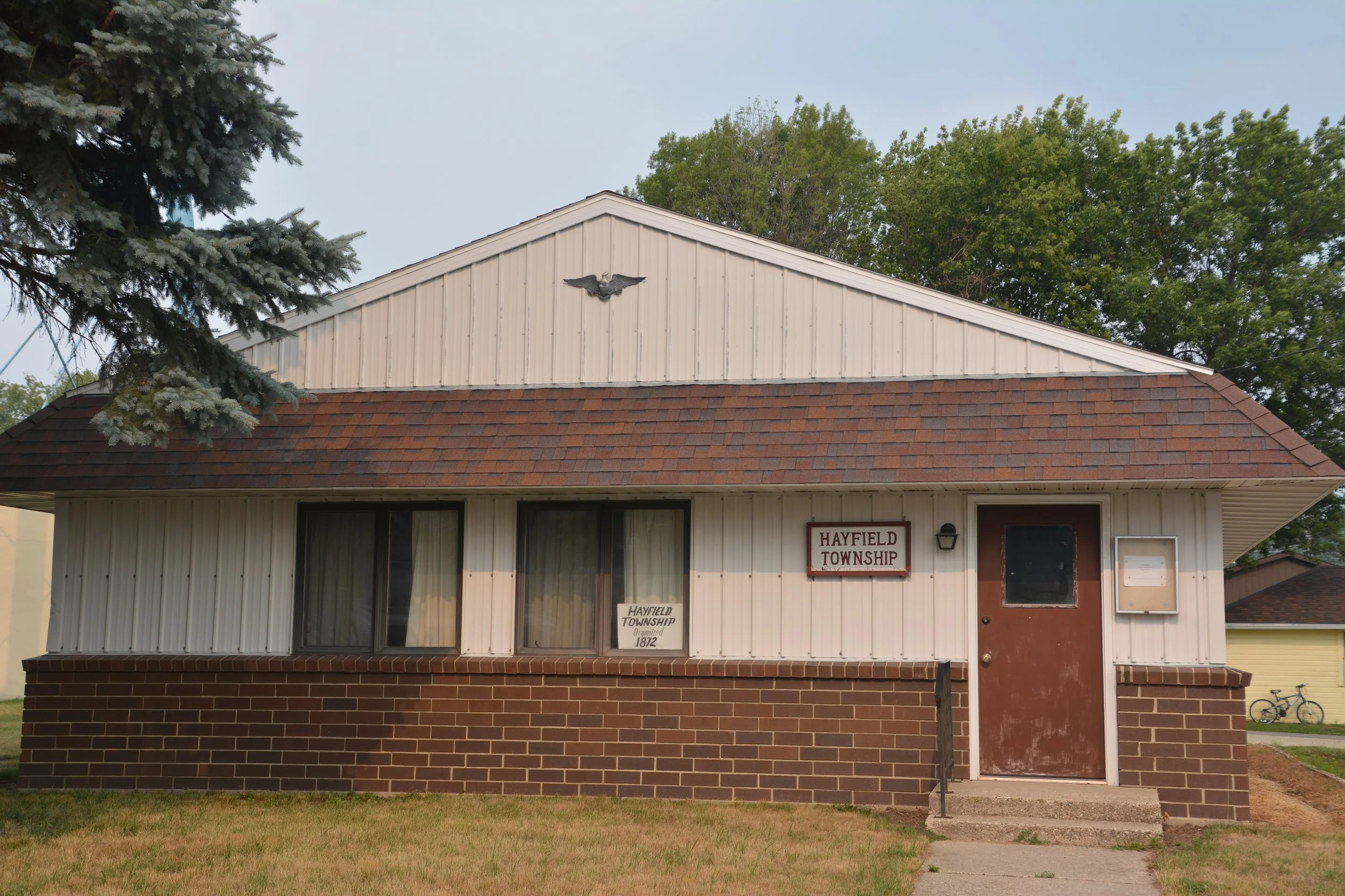 A small government building with beige siding and a brick foundation, featuring an eagle emblem at the front peak and a sign that reads "Hayfield Township". Surrounded by trees and a grassy lawn.