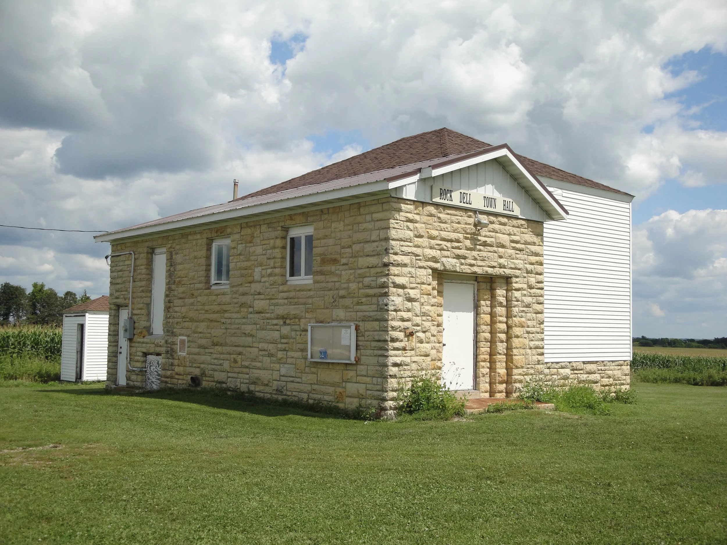 A two-story building with a stone lower level and white siding upper level. The building has a brown shingled roof and a sign that reads 'Rock Dell Town Hall.' The sky above is partly cloudy.