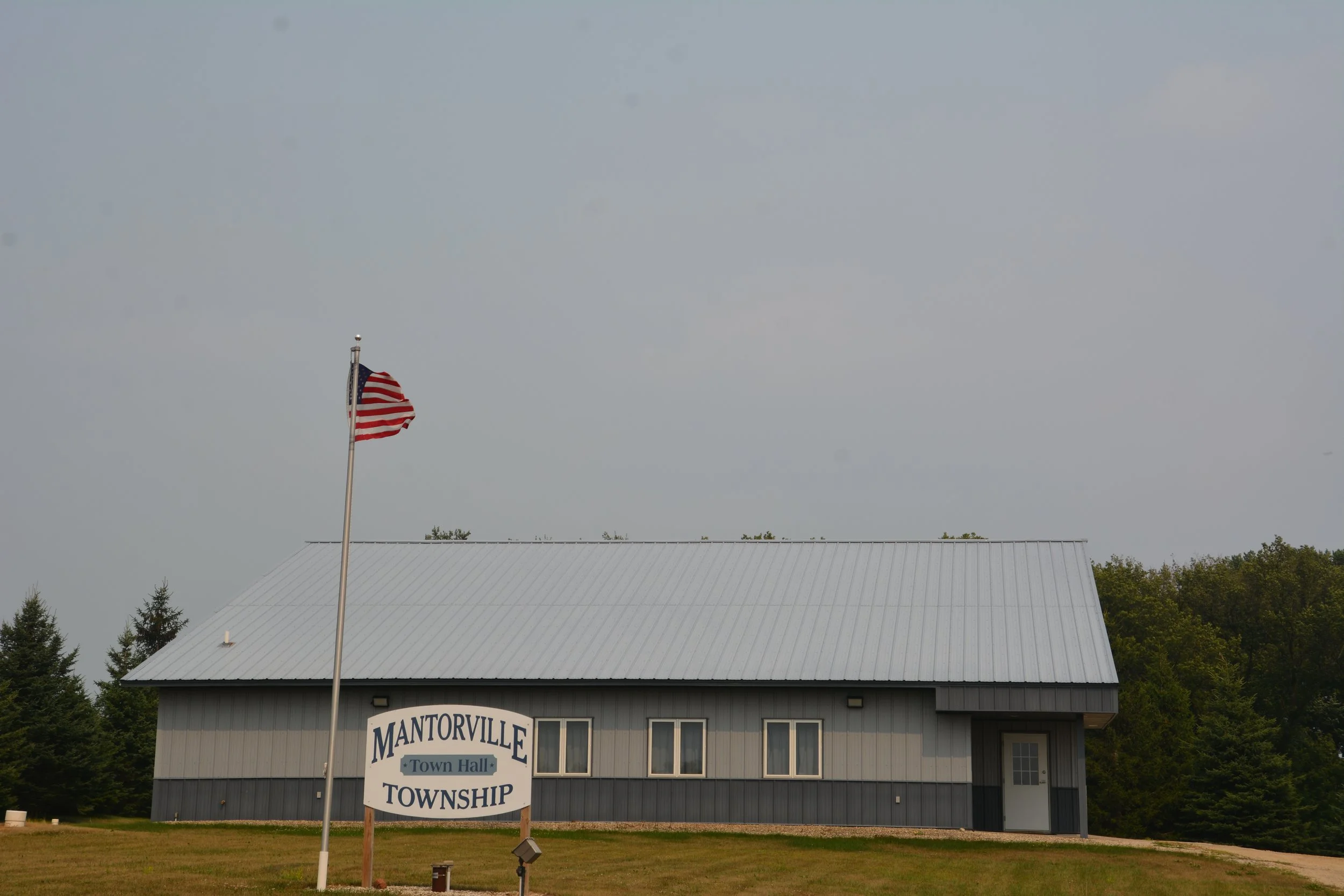 American flag flying on a flagpole in front of a building with a sign that reads 'MANTOVILLE Town Hall TOWNSHIP' under a cloudy sky.