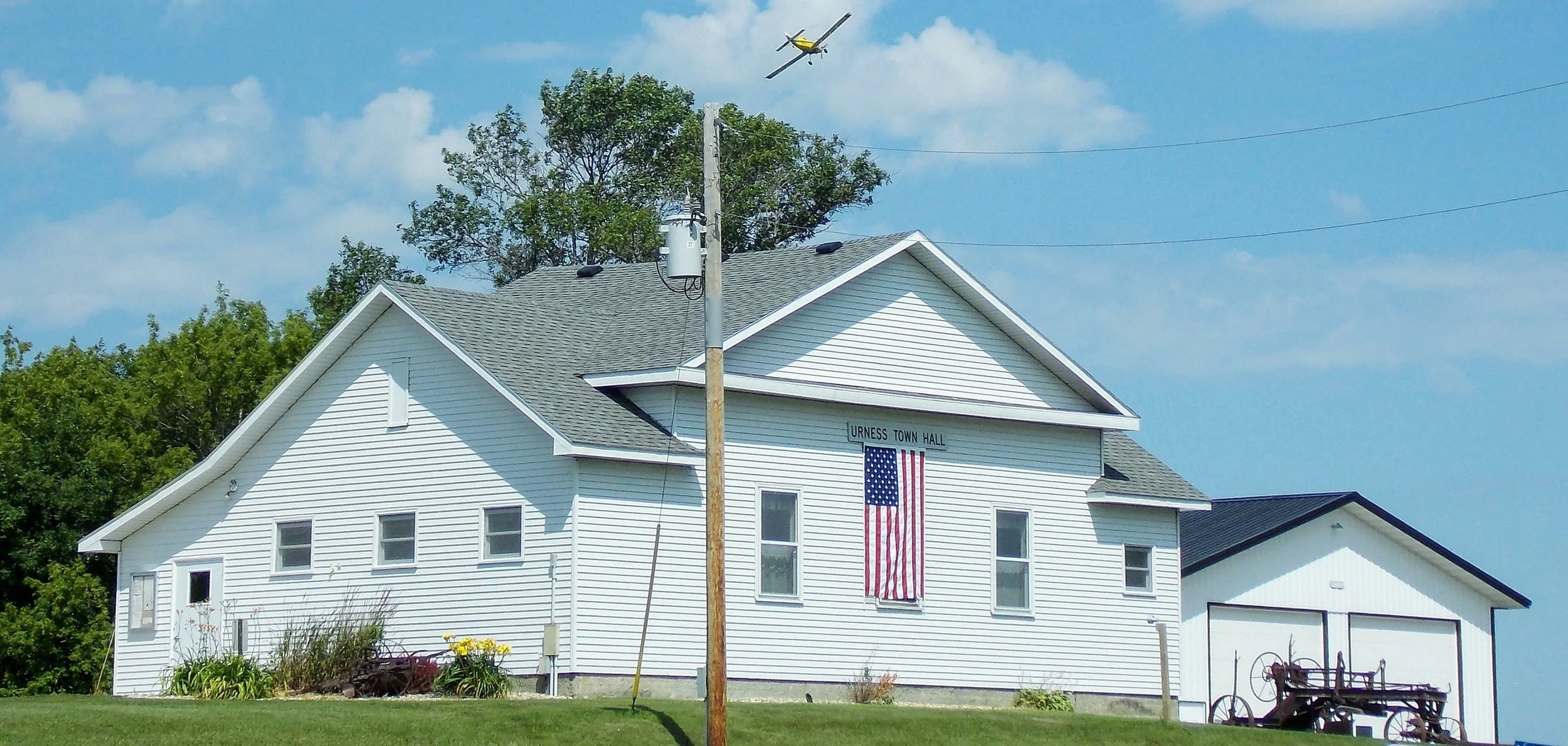 White town hall building with an American flag hanging on the front and the sign 'URNESS TOWN HALL' above it. A yellow airplane is flying in the blue sky overhead. There are trees in the background and a green lawn with some plants and flowers in the