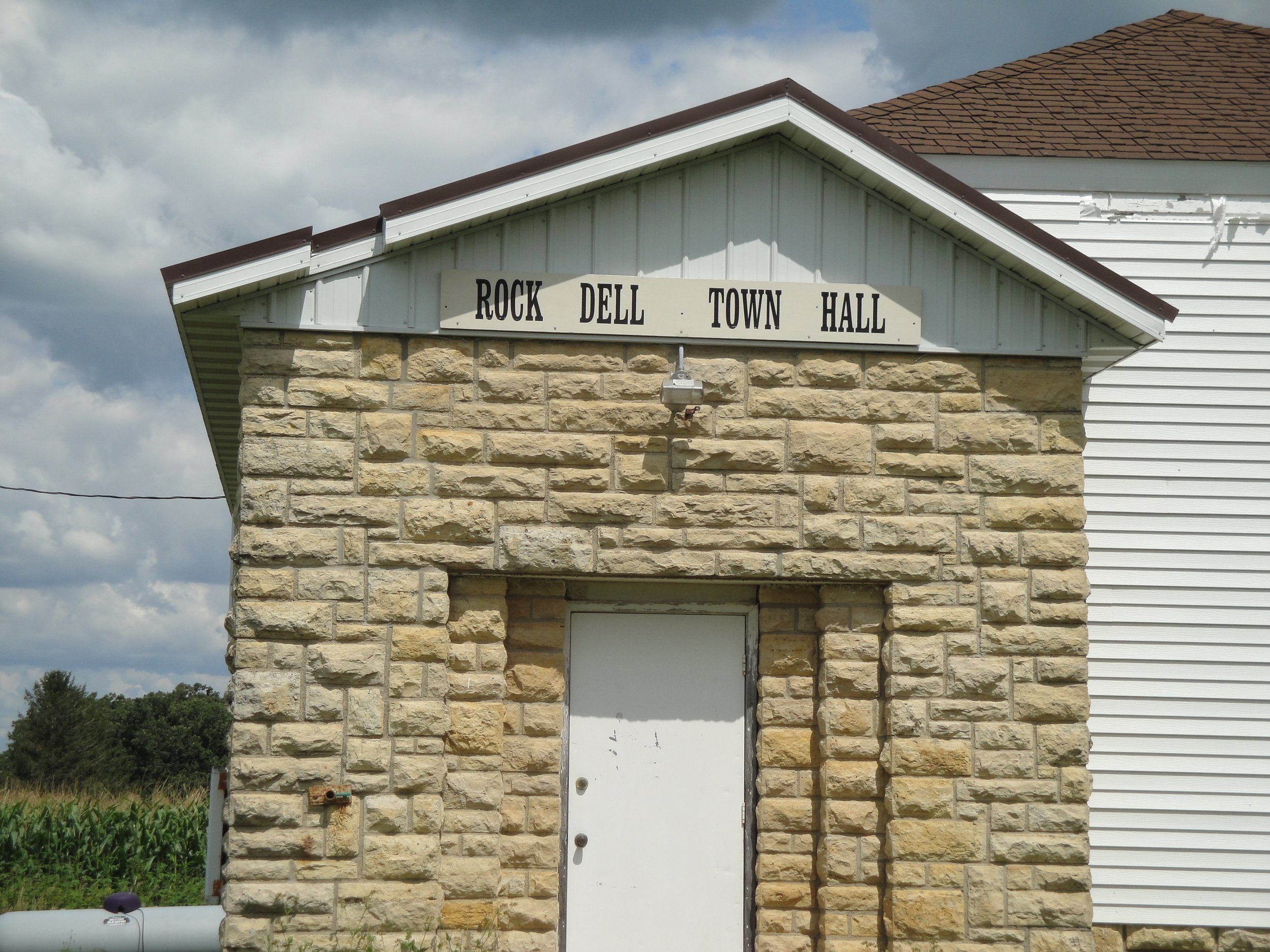 Brick building with a white sign reading 'Rock Dell Town Hall' above the door, under a brown roof with a cloudy sky background.