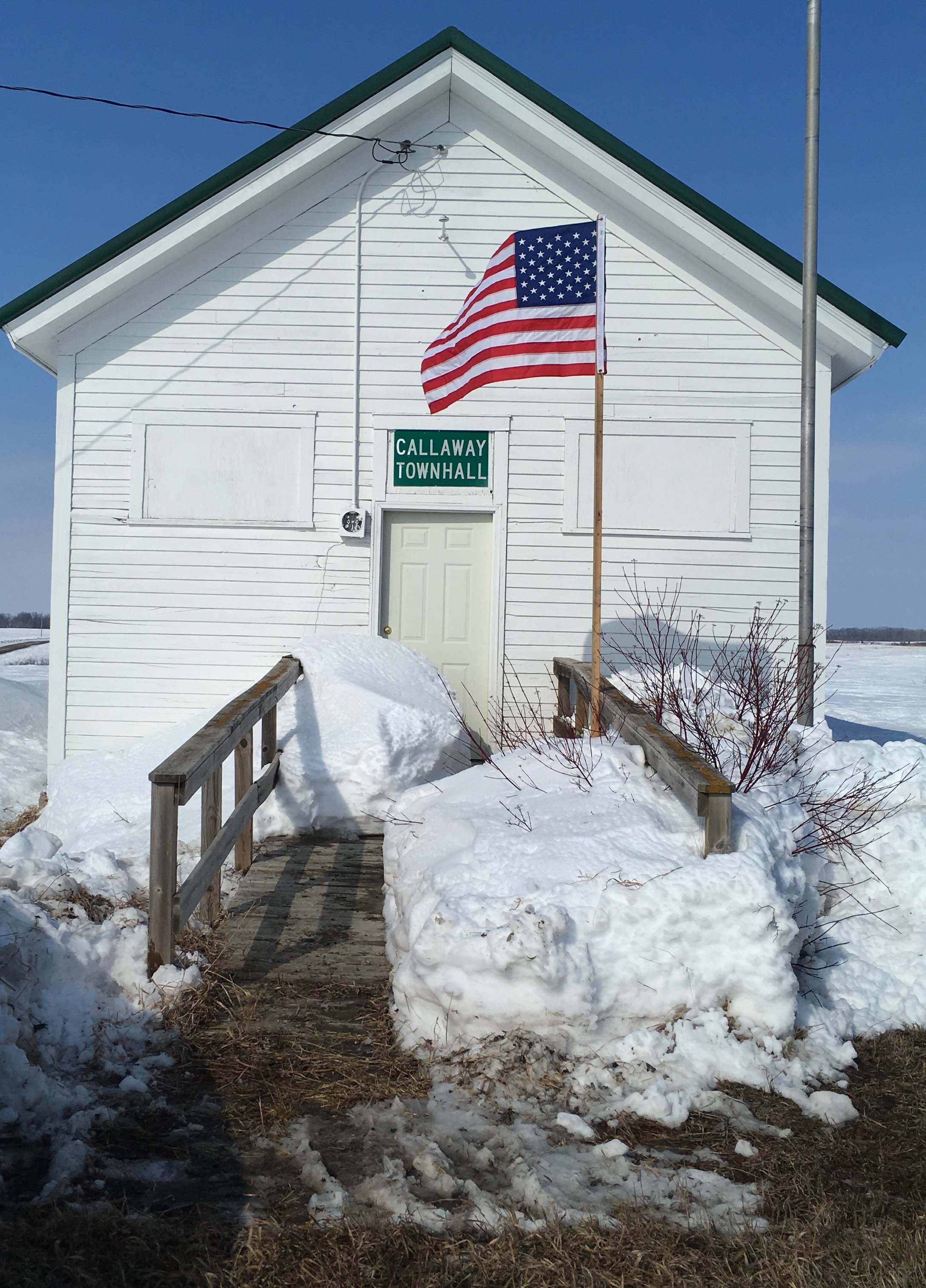A small white wooden building labeled 'Callaway Town Hall' with an American flag flying on a flagpole in front. The ground is covered in snow, and a snow-covered pathway leads up to the entrance.