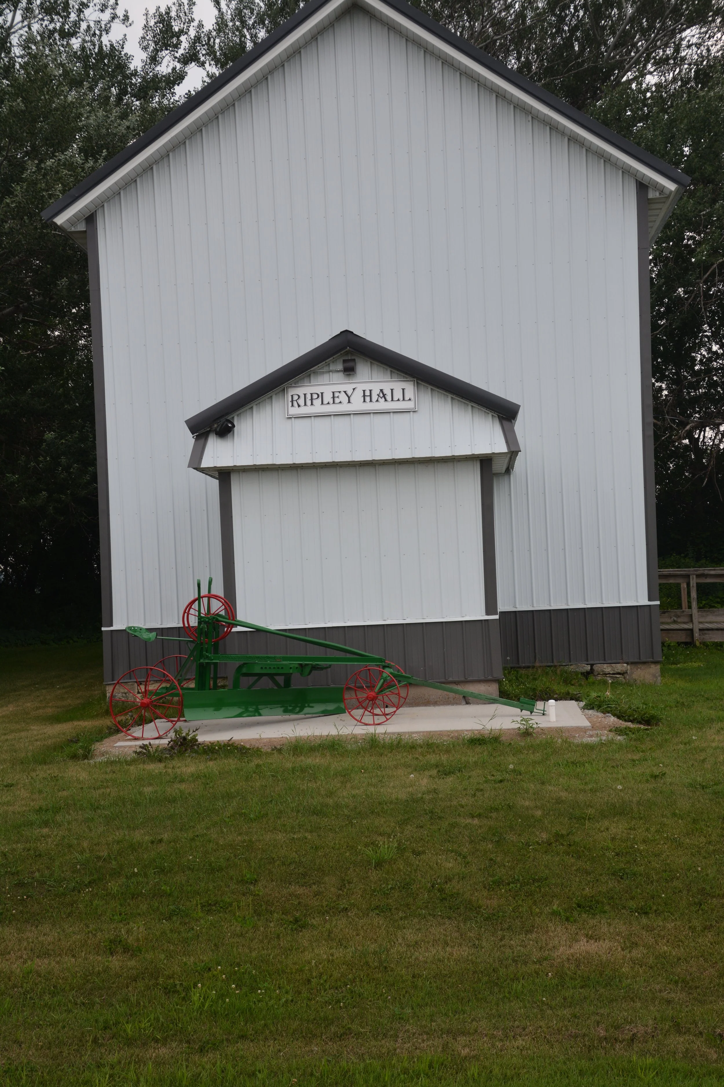 A white wooden building labeled "Ripley Hall" with a gable roof and a small porch. In front, there is a vintage green and red agricultural plow on a concrete slab, surrounded by green grass and trees in the background.