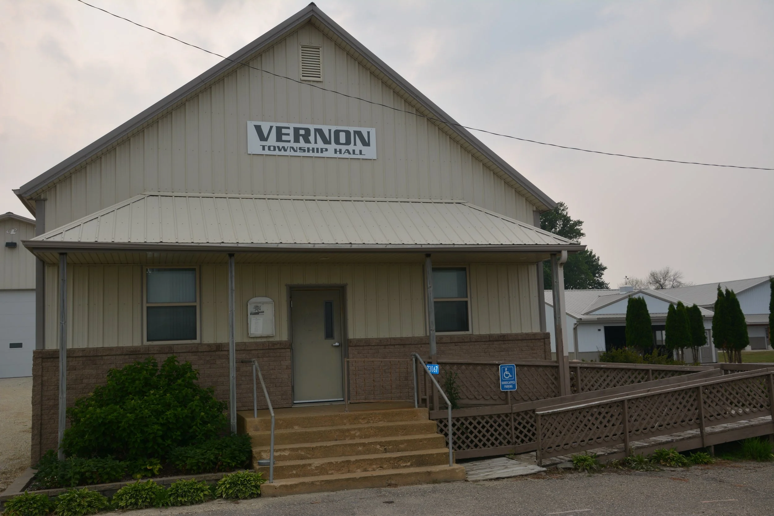 Front view of Vernon Township Hall, a beige two-story building with a metal roof, stairs leading to the entrance, and a sign indicating it is the township hall. There are some bushes and a parking space with handicapped parking signs in front.