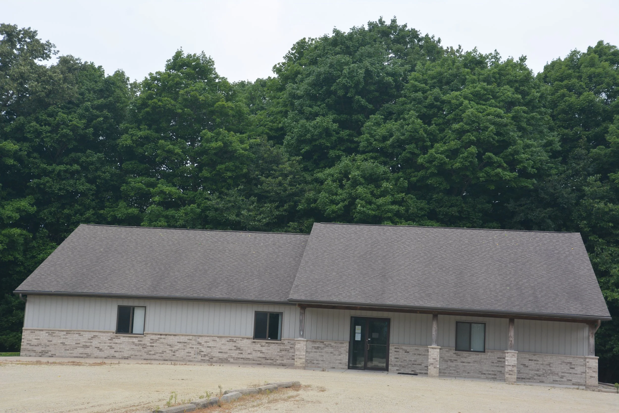 A two-story house with a gray shingle roof, white siding, and gray brick accents at the base, situated in front of large green trees under a cloudy sky.