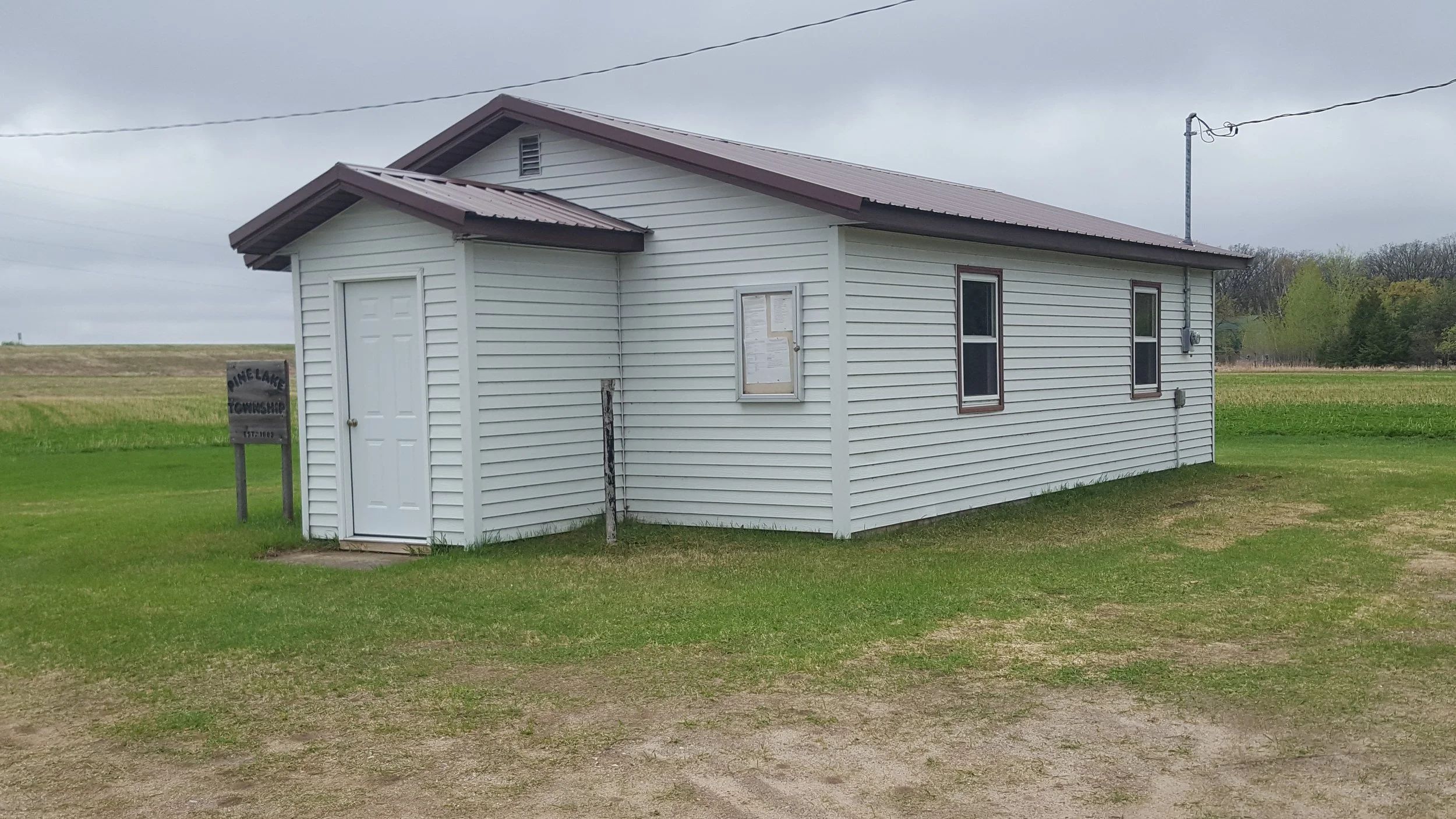 A white building with a metal roof in a grassy open area on a cloudy day. The building has two small windows, a door, and a notice or bulletin board on the front wall. There are electric wires overhead and a sign on the left side indicating it is the