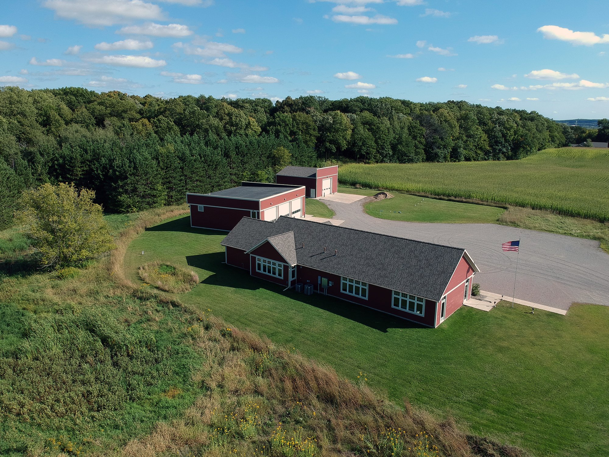Aerial view of a rural landscape featuring two red barns, a grassy yard, a driveway, a flagpole with an American flag, dense green trees, cultivated fields, and a partly cloudy blue sky.