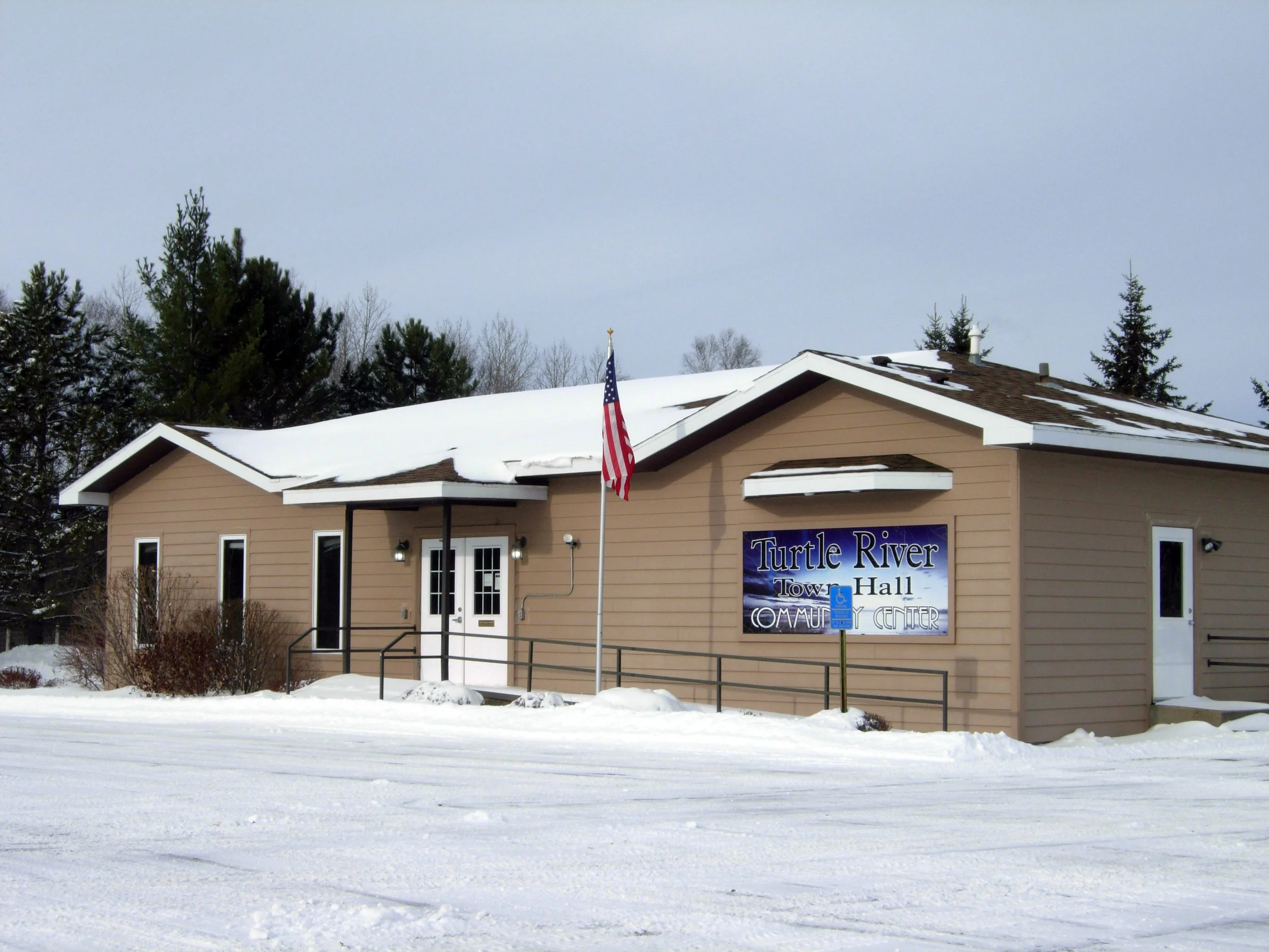 A beige building with a sign that reads 'Turtle River Town Hall Community Center' in a snowy landscape with trees in the background. An American flag is in front of the building and the roof has patches of snow.