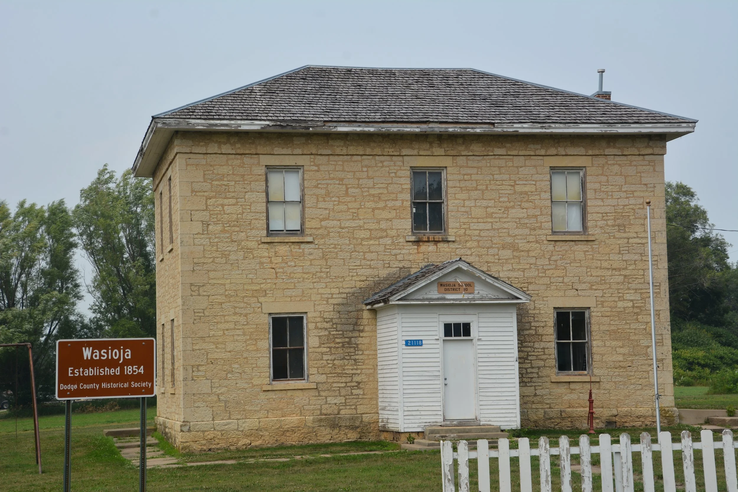 Historic stone building with a small white addition, multiple windows, and a sign indicating it is part of the Dodge County Historical Society, located in Wasioja established in 1854.